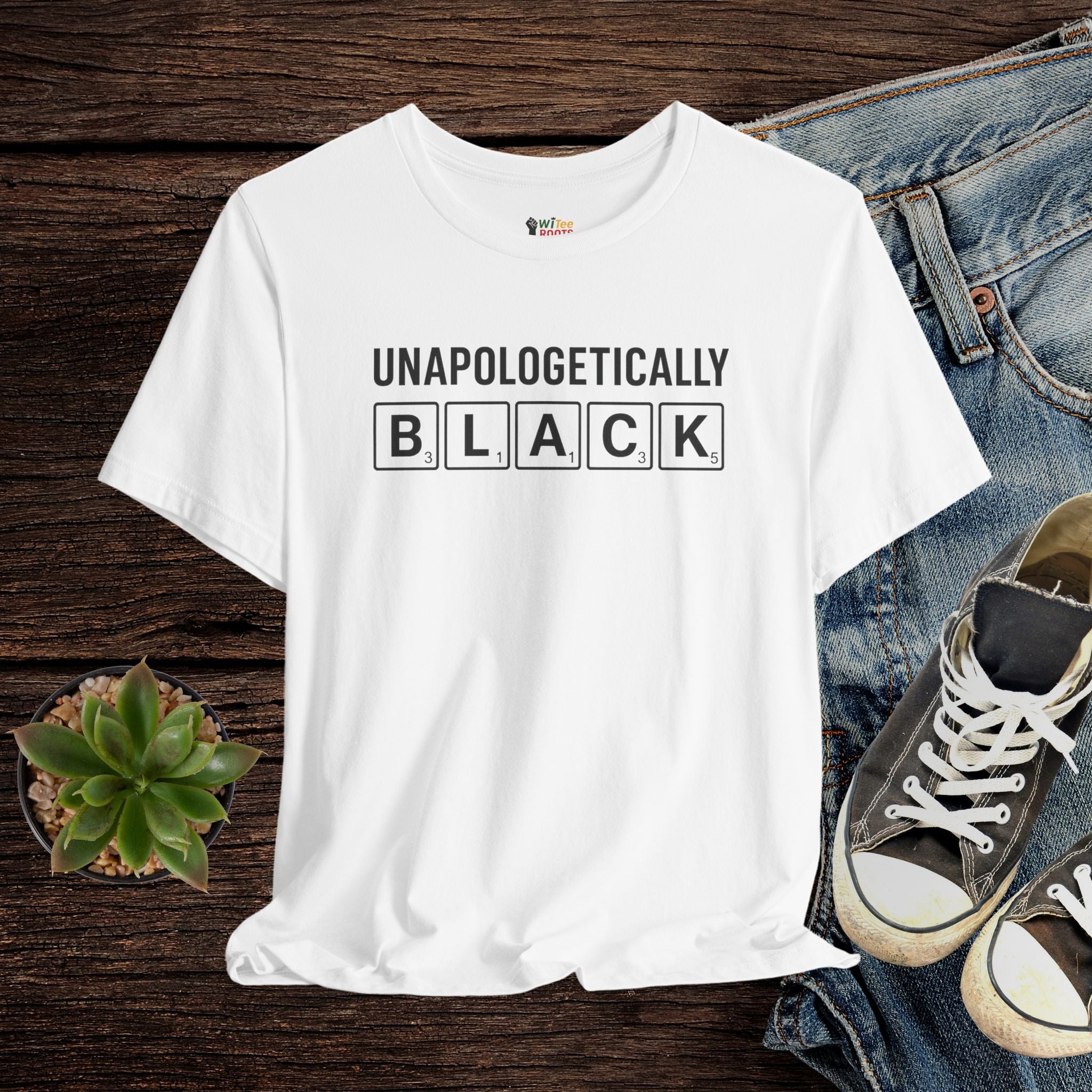 White t-shirt with 'UNAPOLOGETICALLY BLACK' text on a wooden surface with jeans and sneakers.