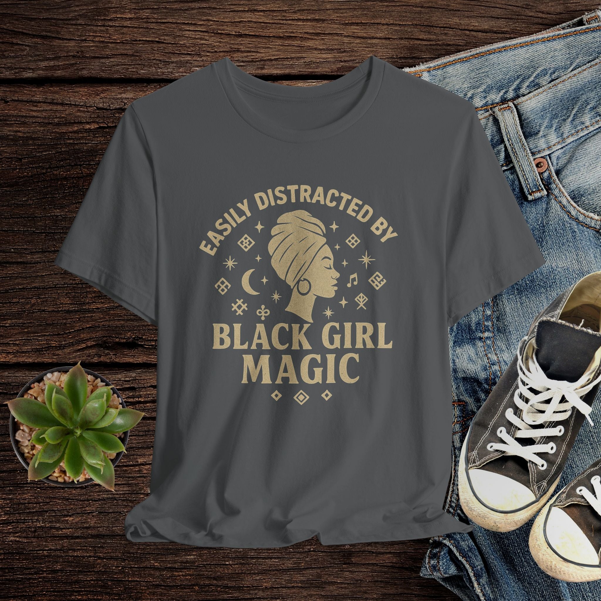 Gray t-shirt with 'Easily Distracted by Black Girl Magic' design on a wooden surface with jeans and sneakers.