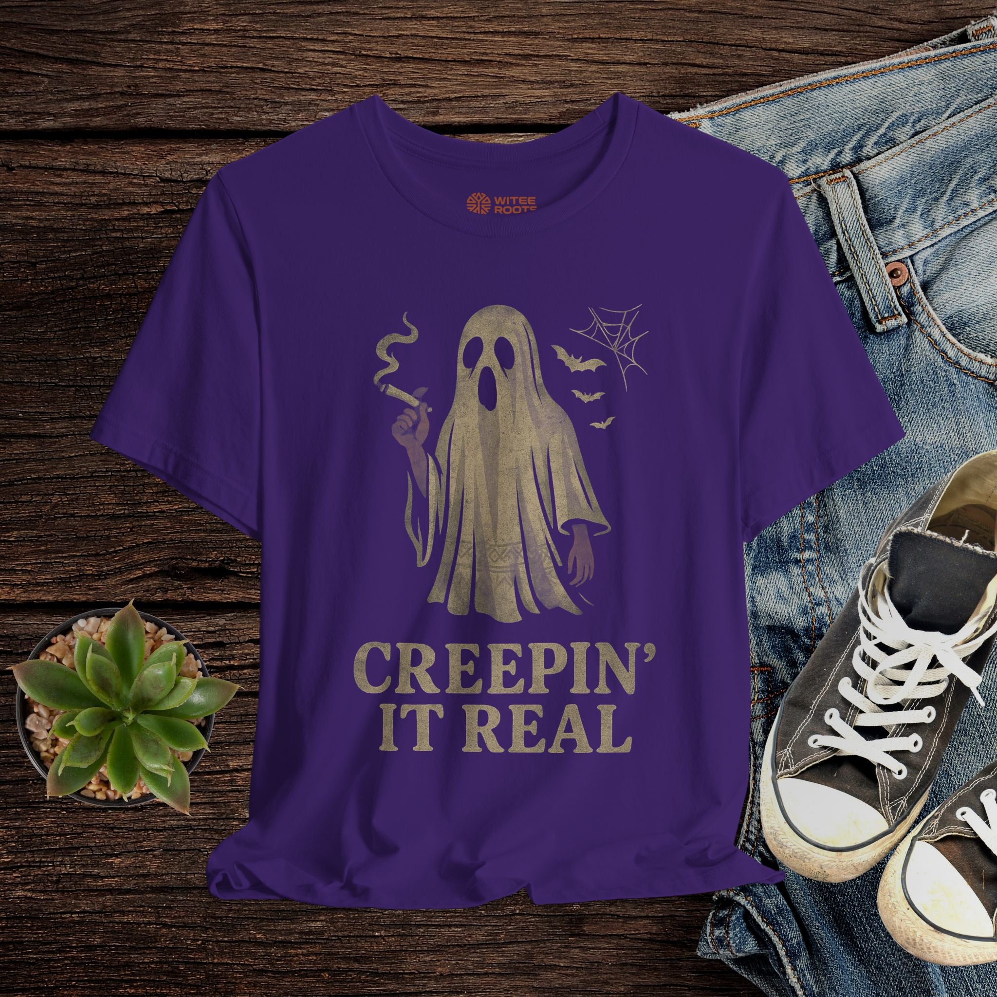 Purple t-shirt with ghost graphic and text 'Creepin' It Real' on a wooden surface with jeans and sneakers.