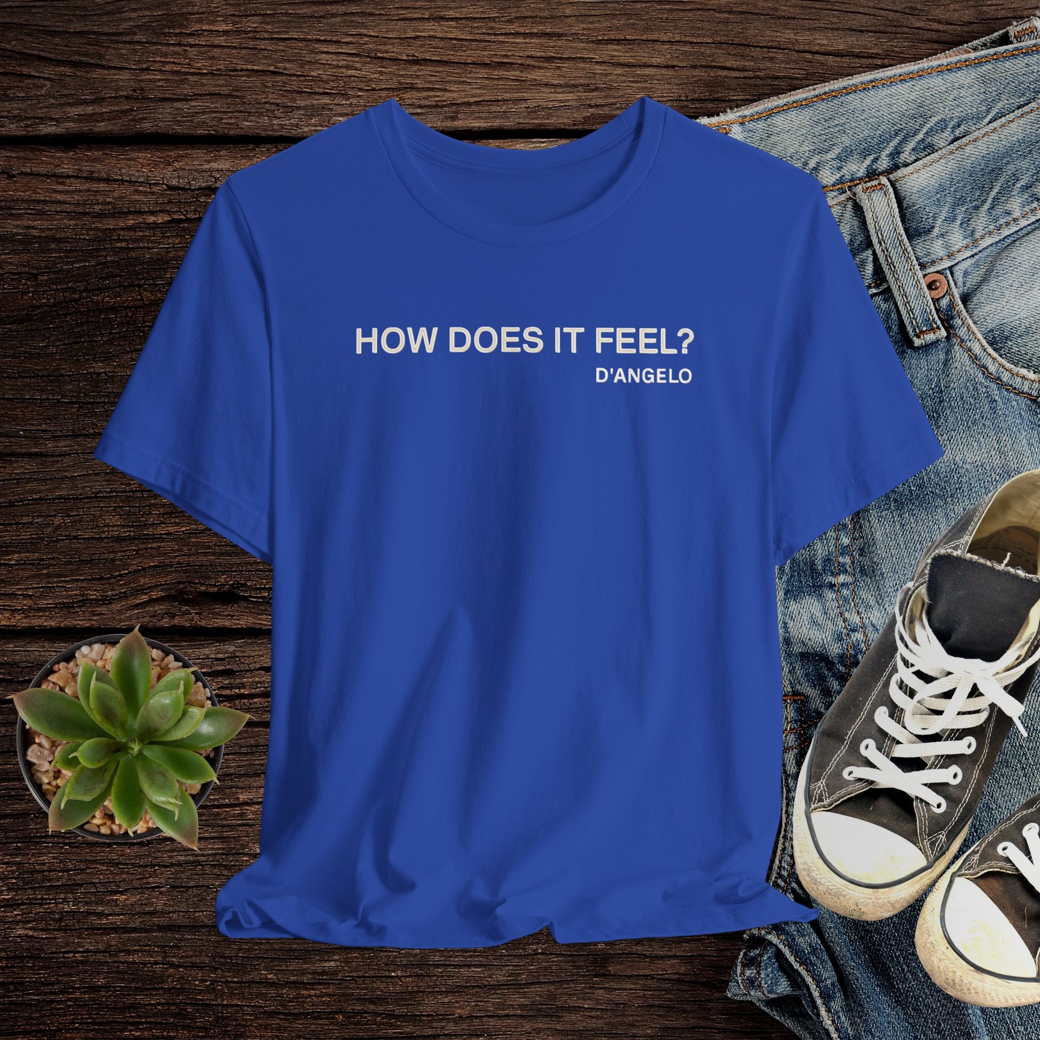 Blue t-shirt with text 'HOW DOES IT FEEL? D'ANGELO' on a wooden surface with jeans and sneakers.