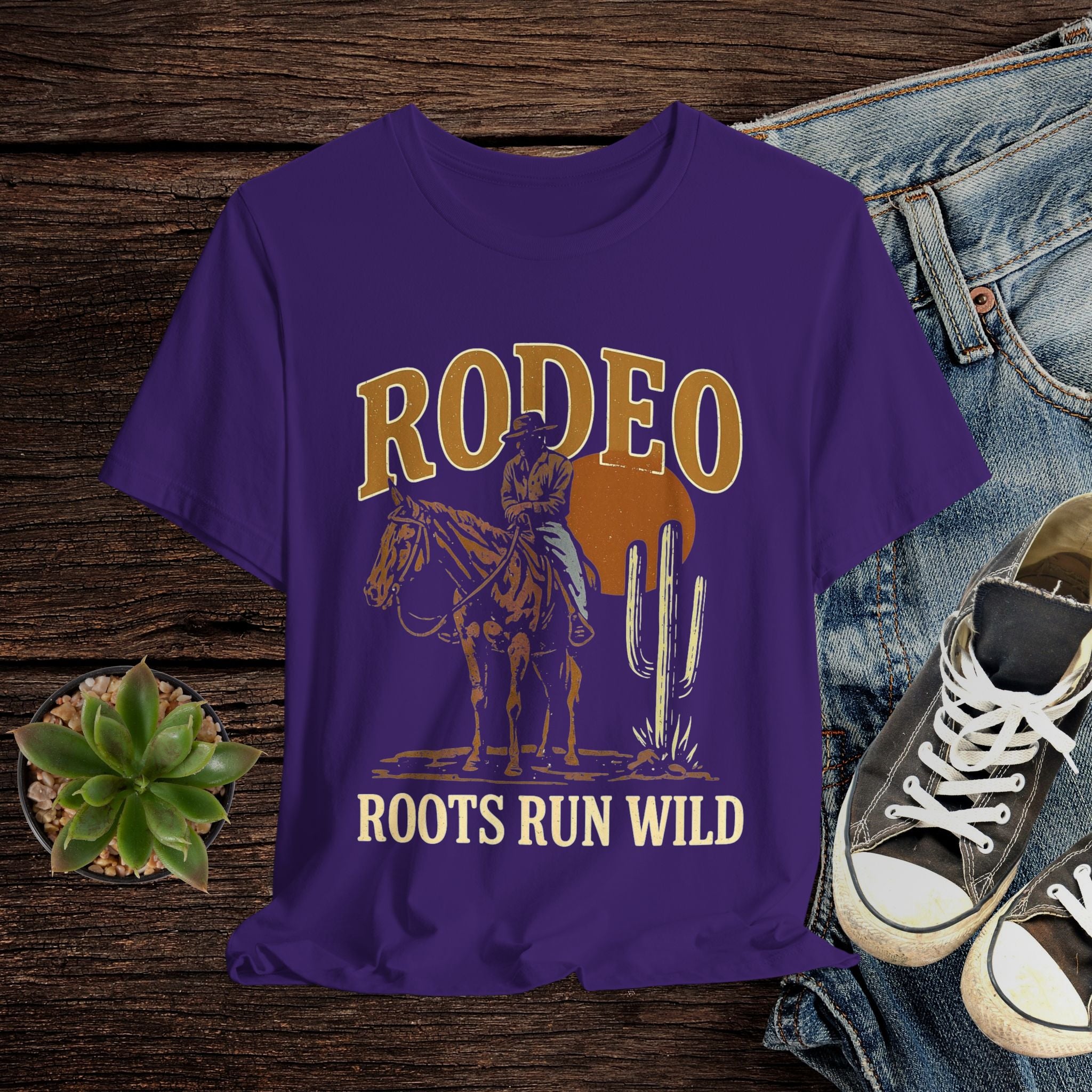 Purple t-shirt with cowboy graphic and 'Rodeo Roots Run Wild' text on a wooden surface with jeans and sneakers.