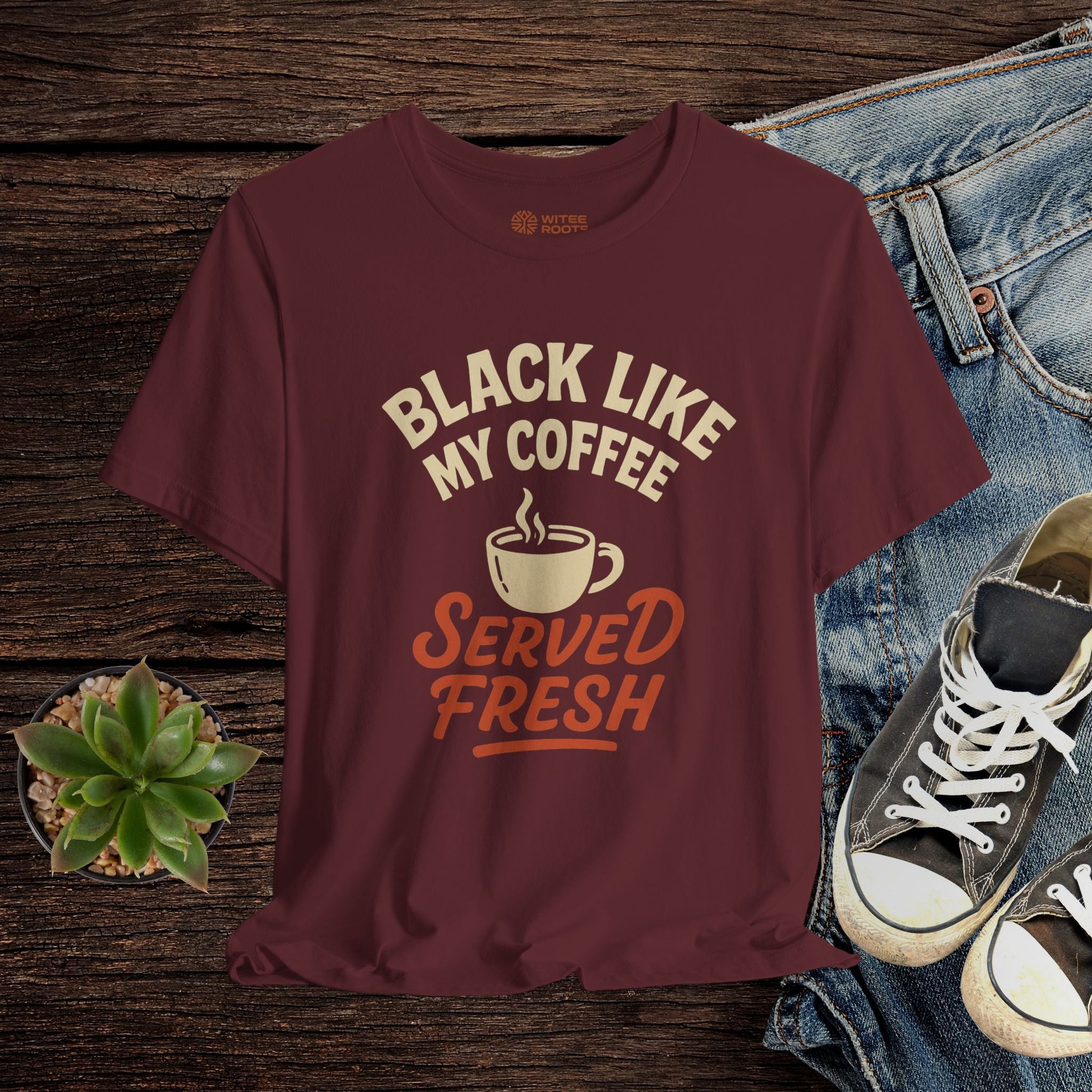 Maroon t-shirt with coffee-themed text on a wooden surface with jeans and sneakers.