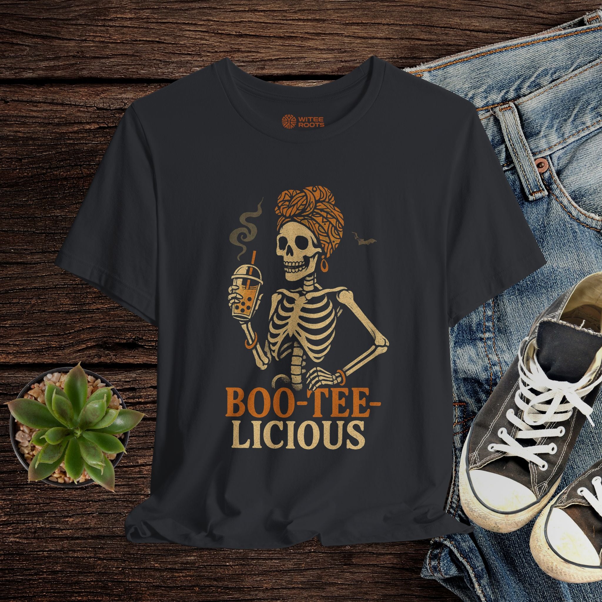 Black t-shirt with skeleton design and 'BOO-TEE-LICIOUS' text on a wooden surface with jeans and sneakers.