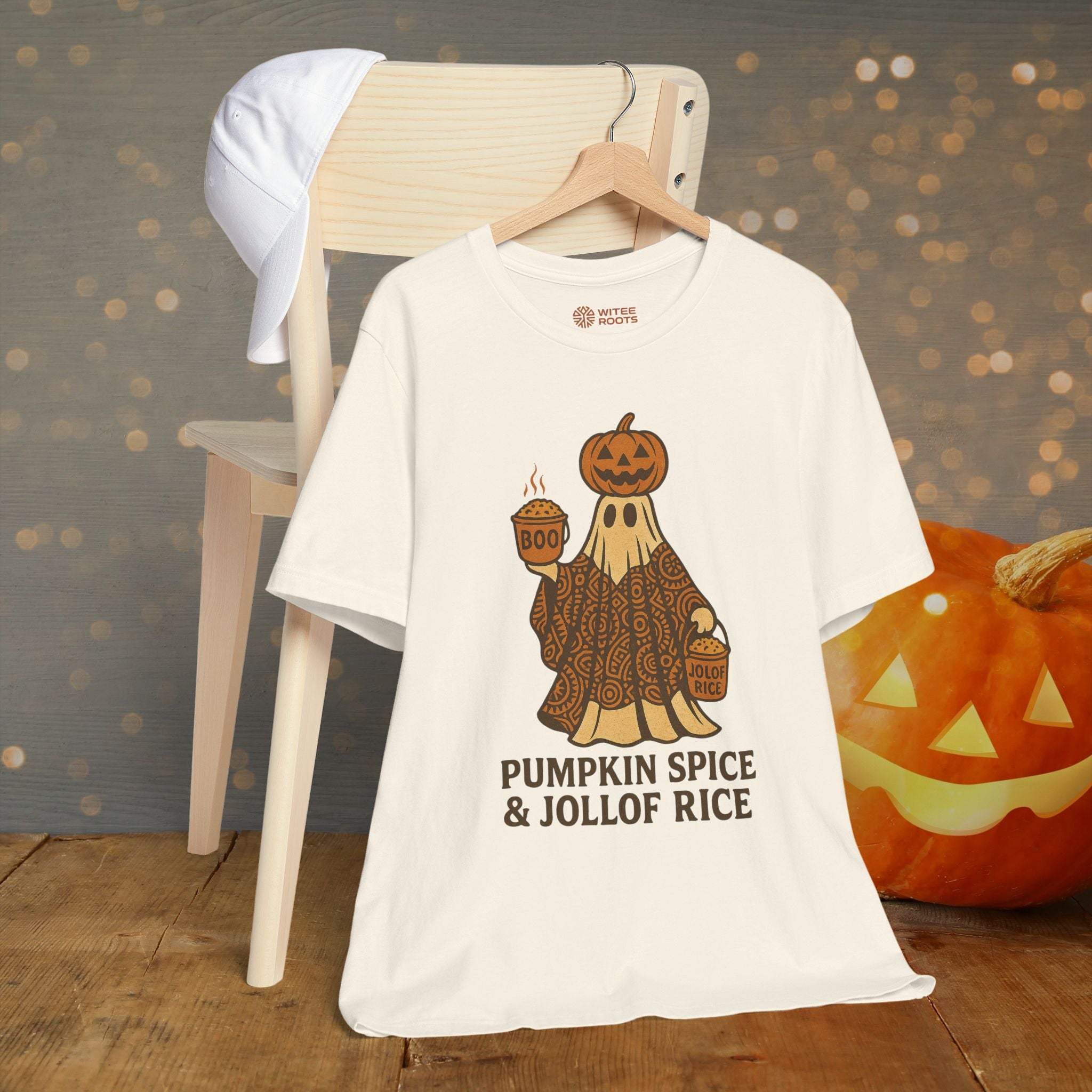 Beige t-shirt with a ghost holding pumpkins and text 'Pumpkin Spice & Jollof Rice' on a wooden surface with a blurred background.
