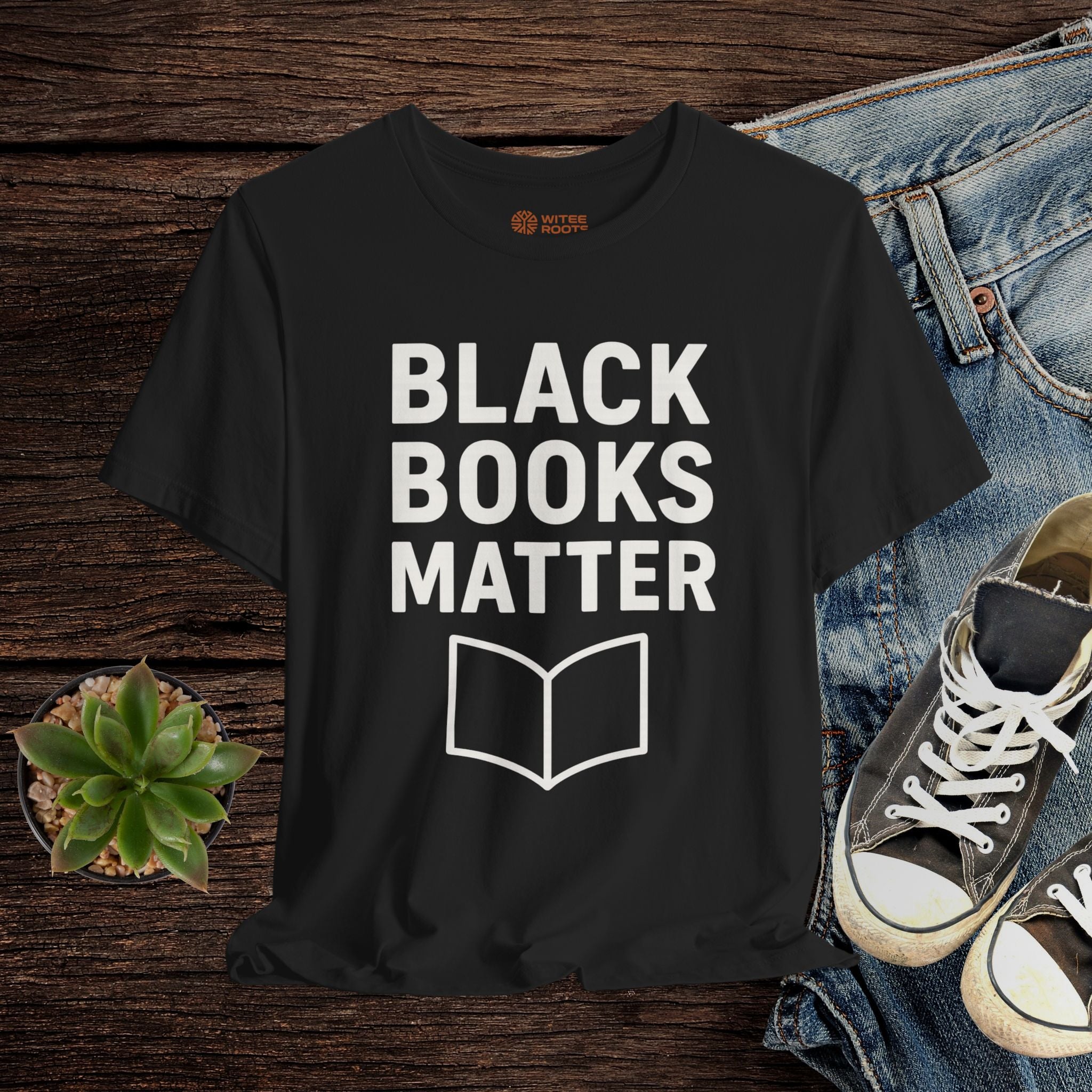 Black t-shirt with 'BLACK BOOKS MATTER' text and book icon on a wooden surface with jeans and sneakers.