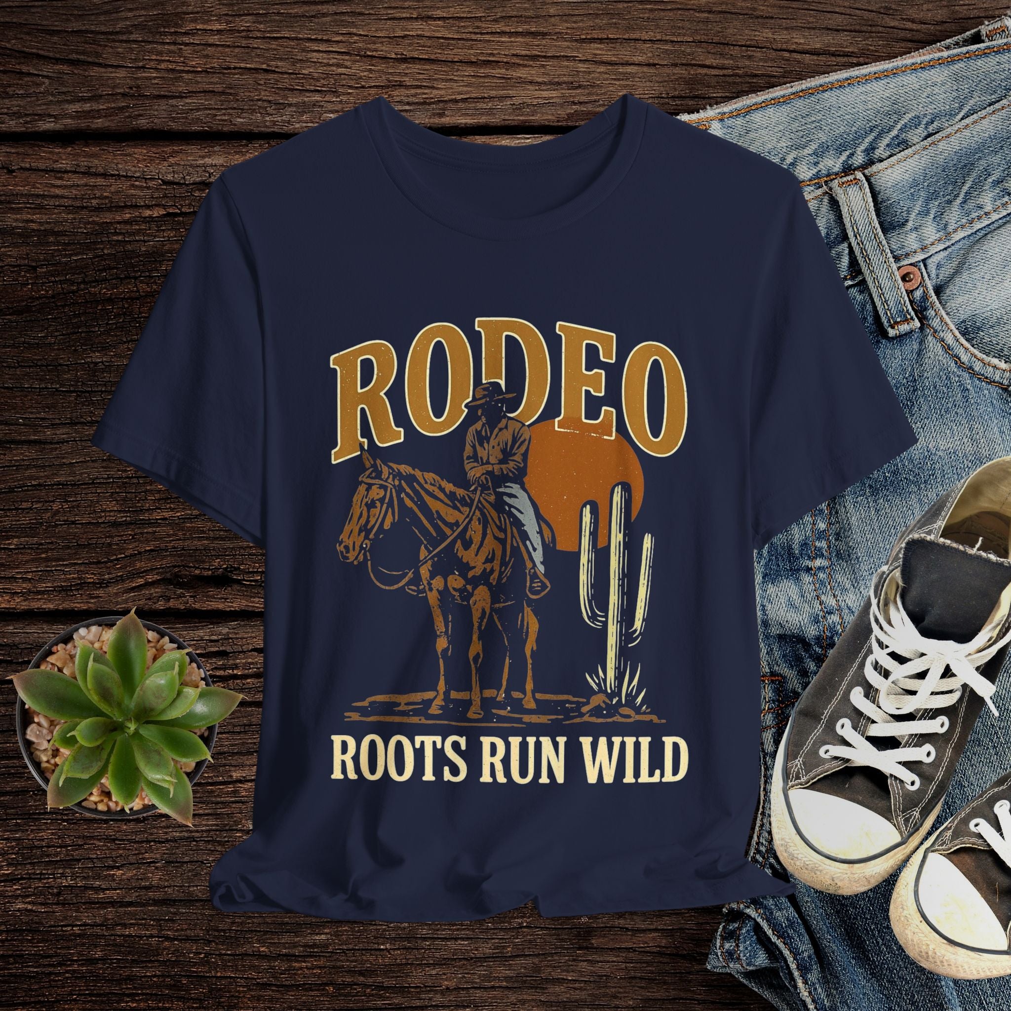 Navy blue t-shirt with cowboy graphic and 'Rodeo Roots Run Wild' text on a wooden surface with jeans and sneakers.