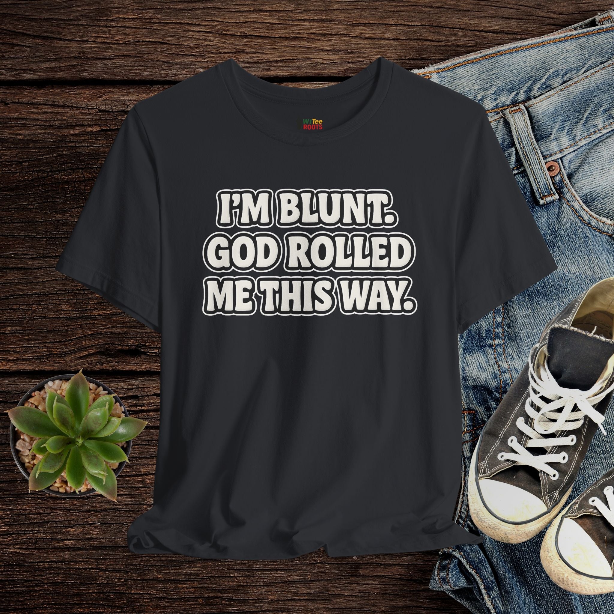 Black t-shirt with text 'I'M BLUNT. GOD ROLLED ME THIS WAY.' on a wooden surface with jeans and sneakers.