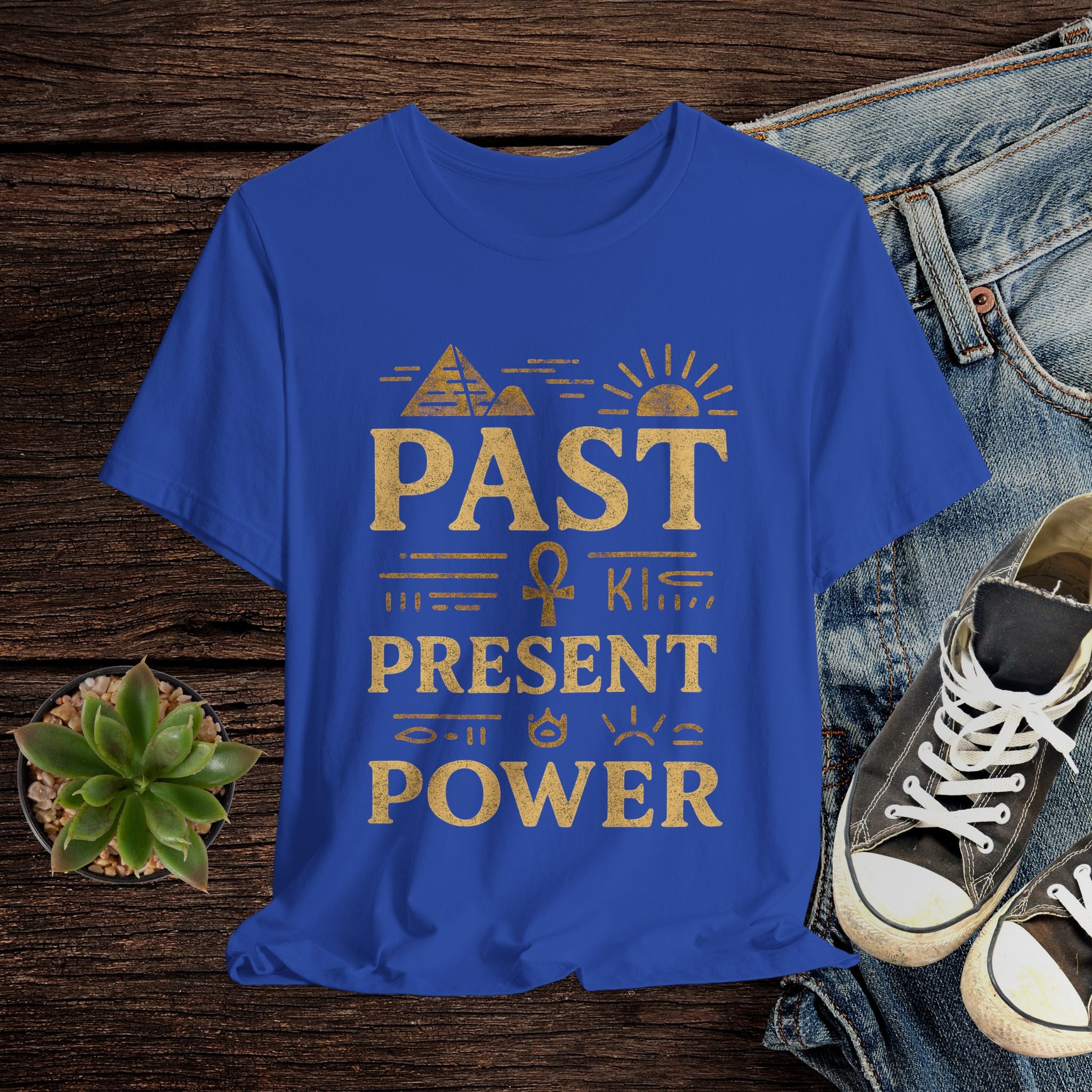 Blue t-shirt with gold text 'Past is Present Power' on a wooden surface with jeans and shoes.