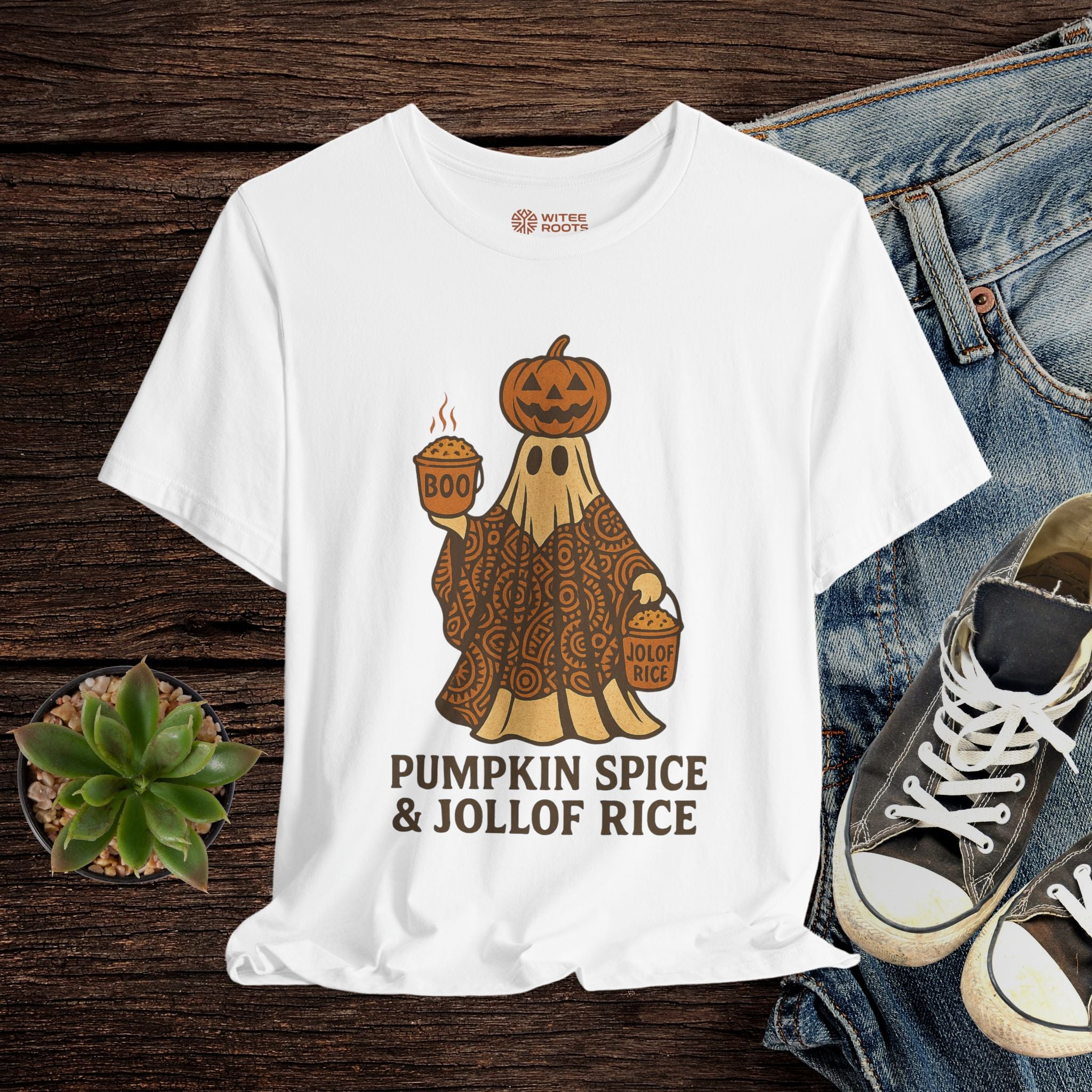 White t-shirt with a graphic design featuring a ghost in a pumpkin costume and text 'Pumpkin Spice & Jollof Rice' on a wooden surface with jeans and sneakers.