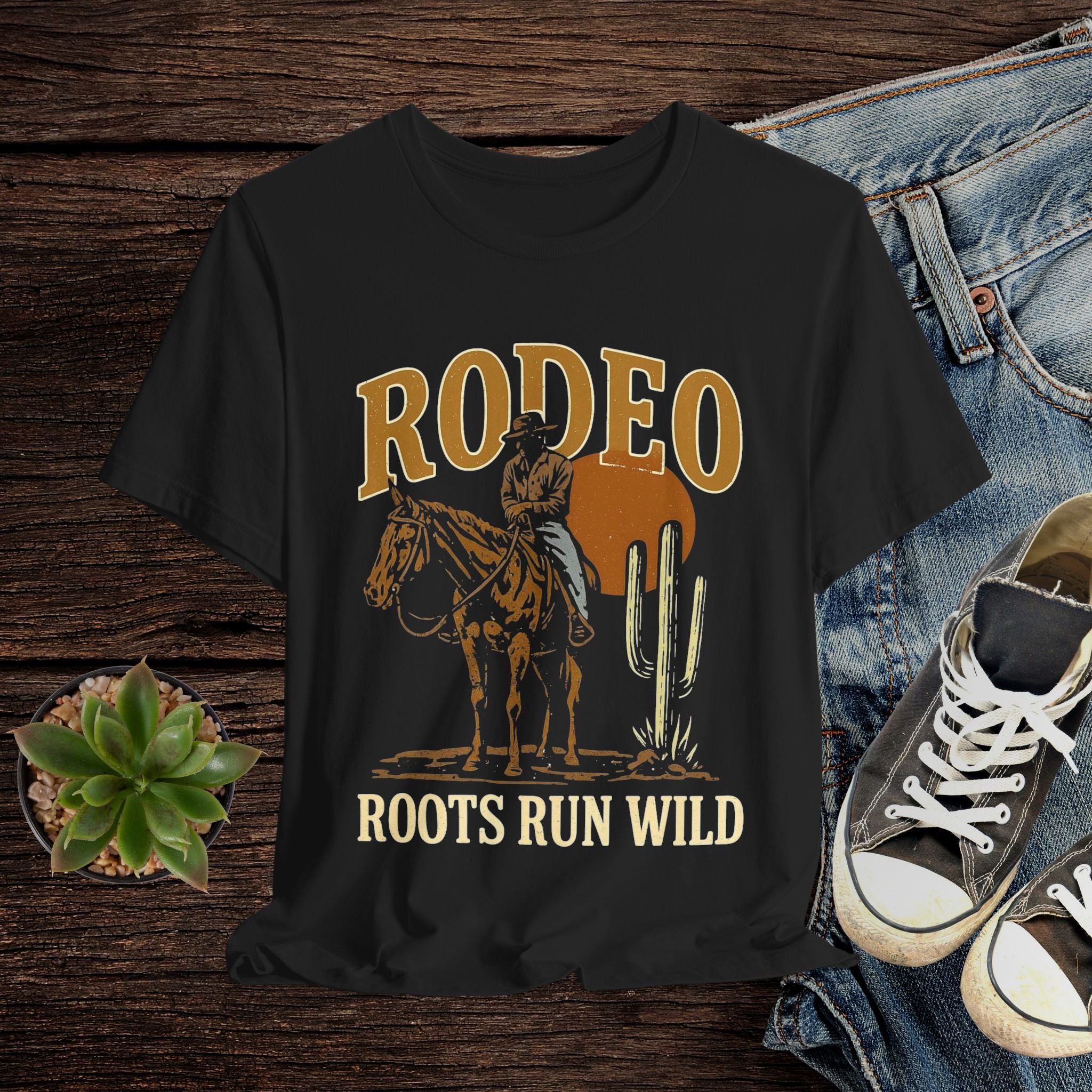 Black t-shirt with cowboy graphic and 'Rodeo Roots Run Wild' text on a wooden surface with jeans and sneakers.