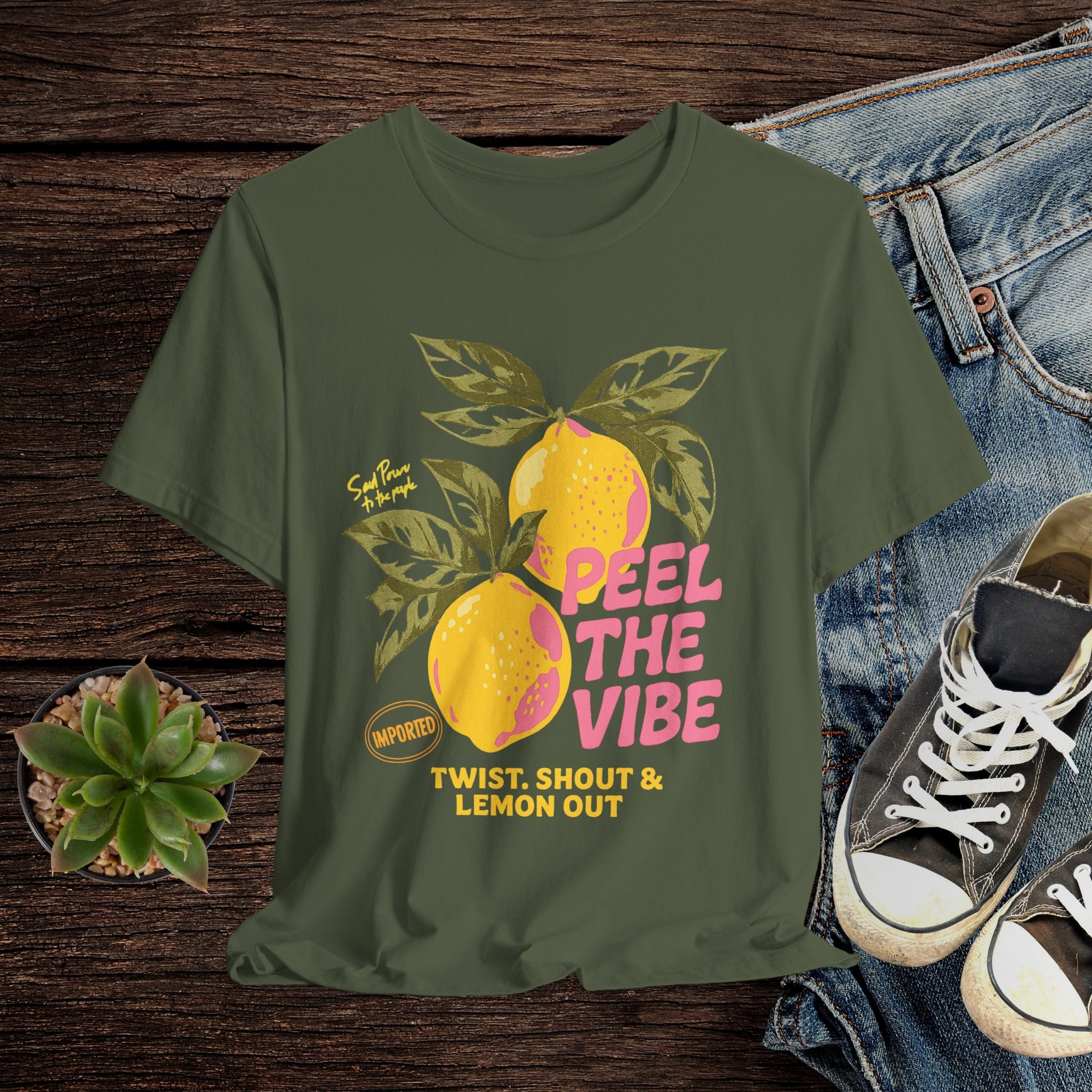 Green t-shirt with lemon graphic and text on a wooden surface with jeans and shoes.