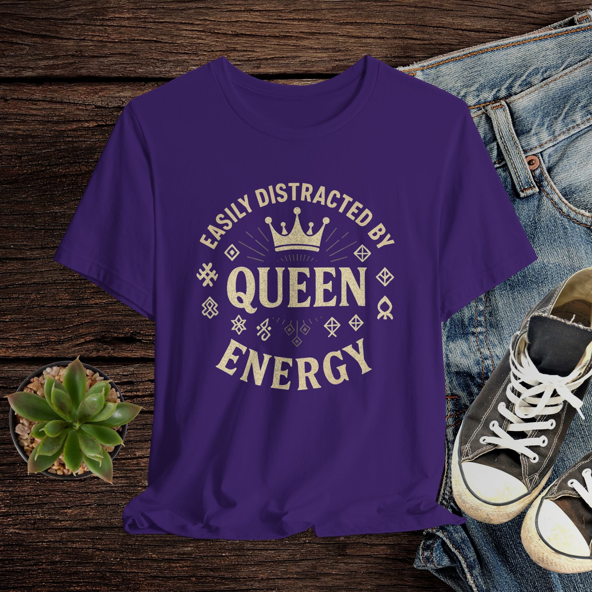 Purple t-shirt with 'Easily Distracted by Queen Energy' text on a wooden surface with jeans and sneakers.