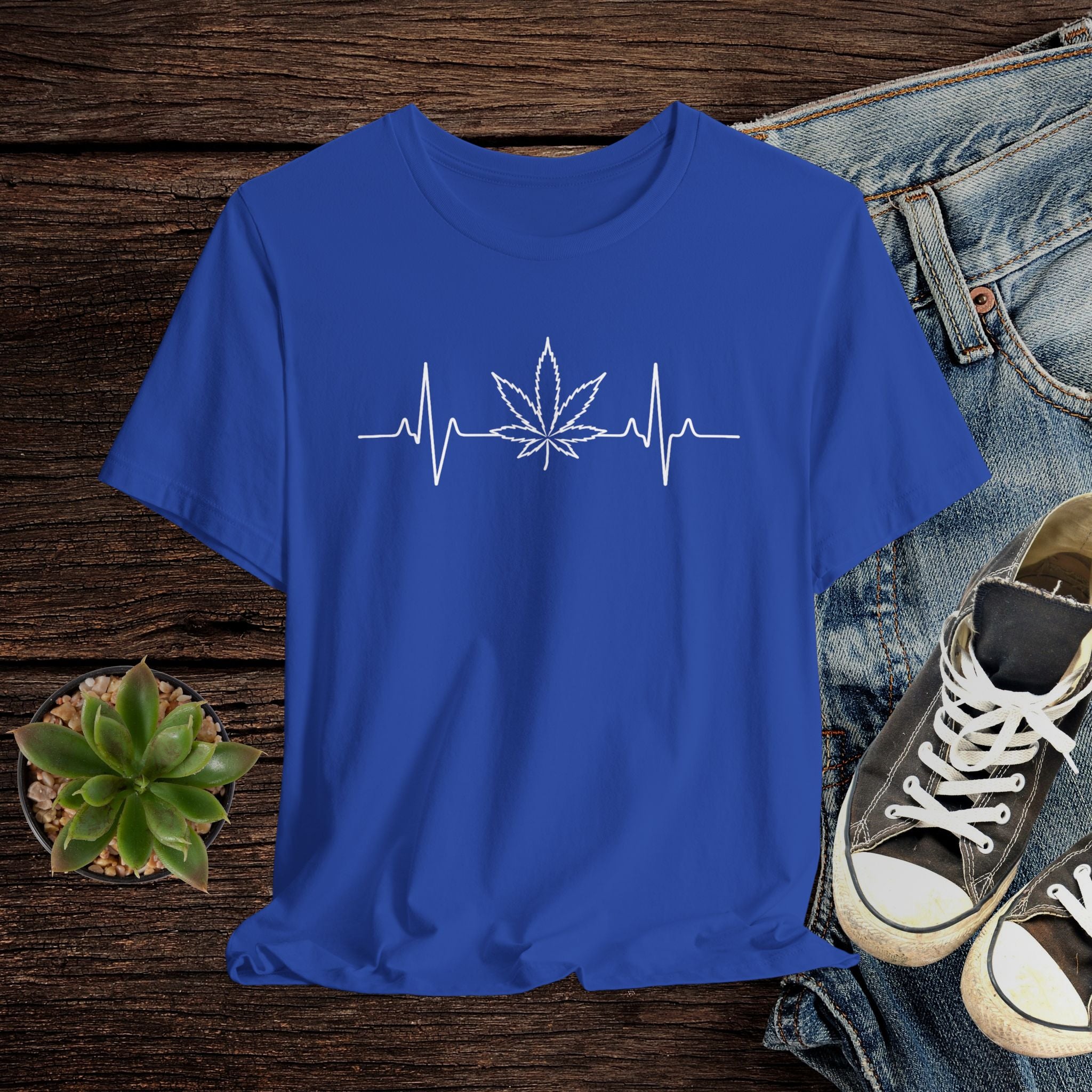 Blue t-shirt with a cannabis leaf and heartbeat design on a wooden surface with jeans and sneakers.