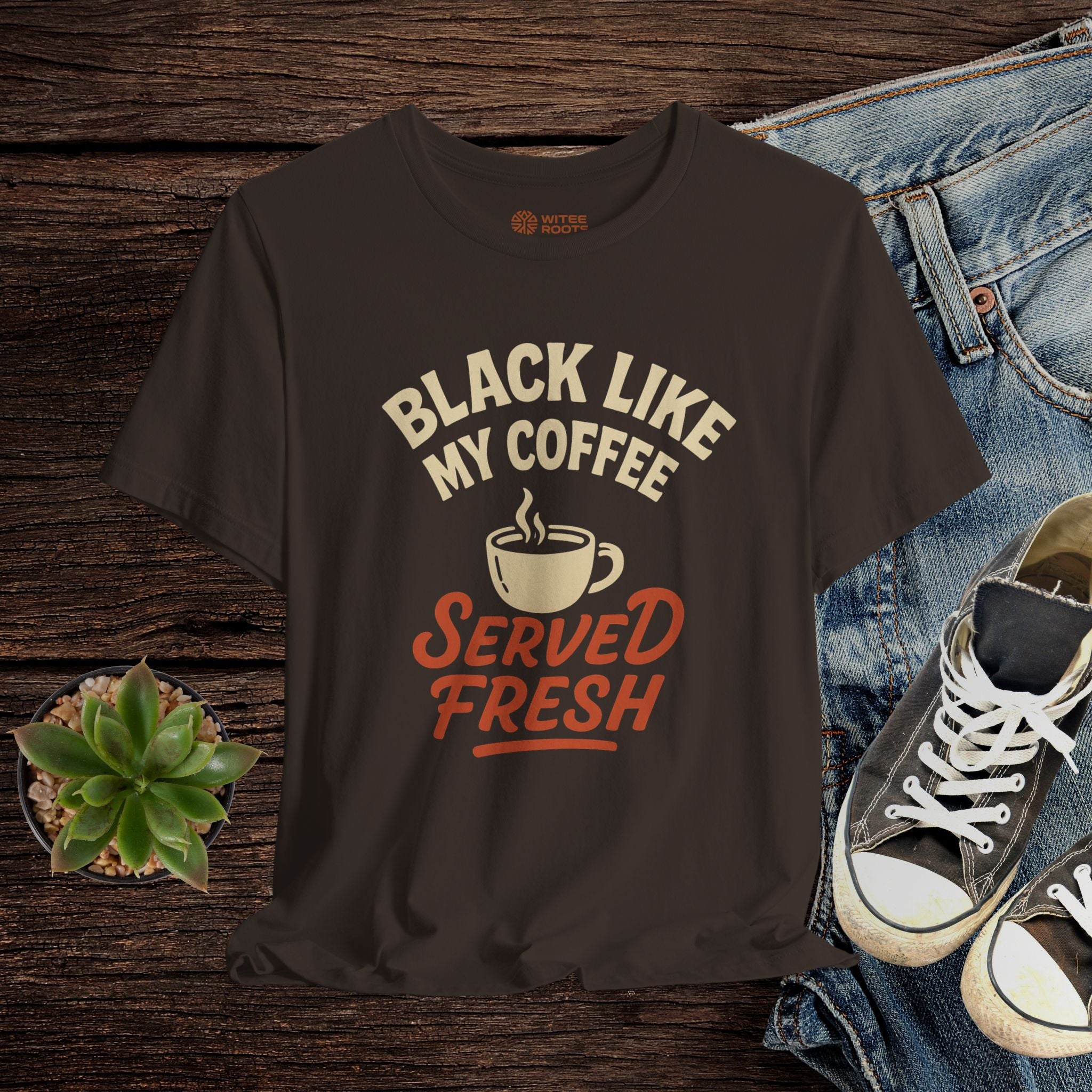Black t-shirt with 'Black Like My Coffee Served Fresh' text and graphic on a wooden surface with jeans and sneakers.