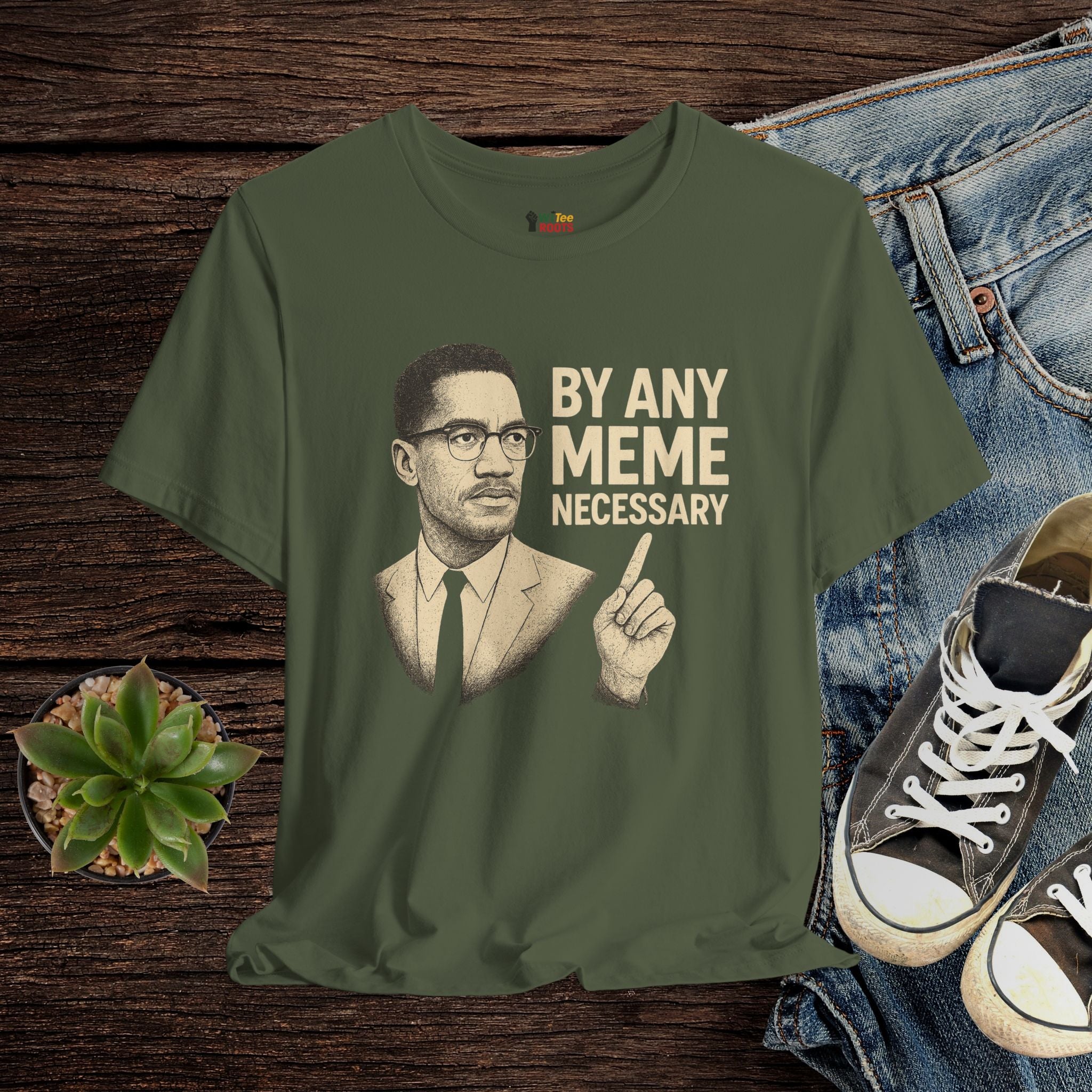 Green t-shirt with a graphic of a man and text 'By Any Meme Necessary' on a wooden surface with jeans and shoes.