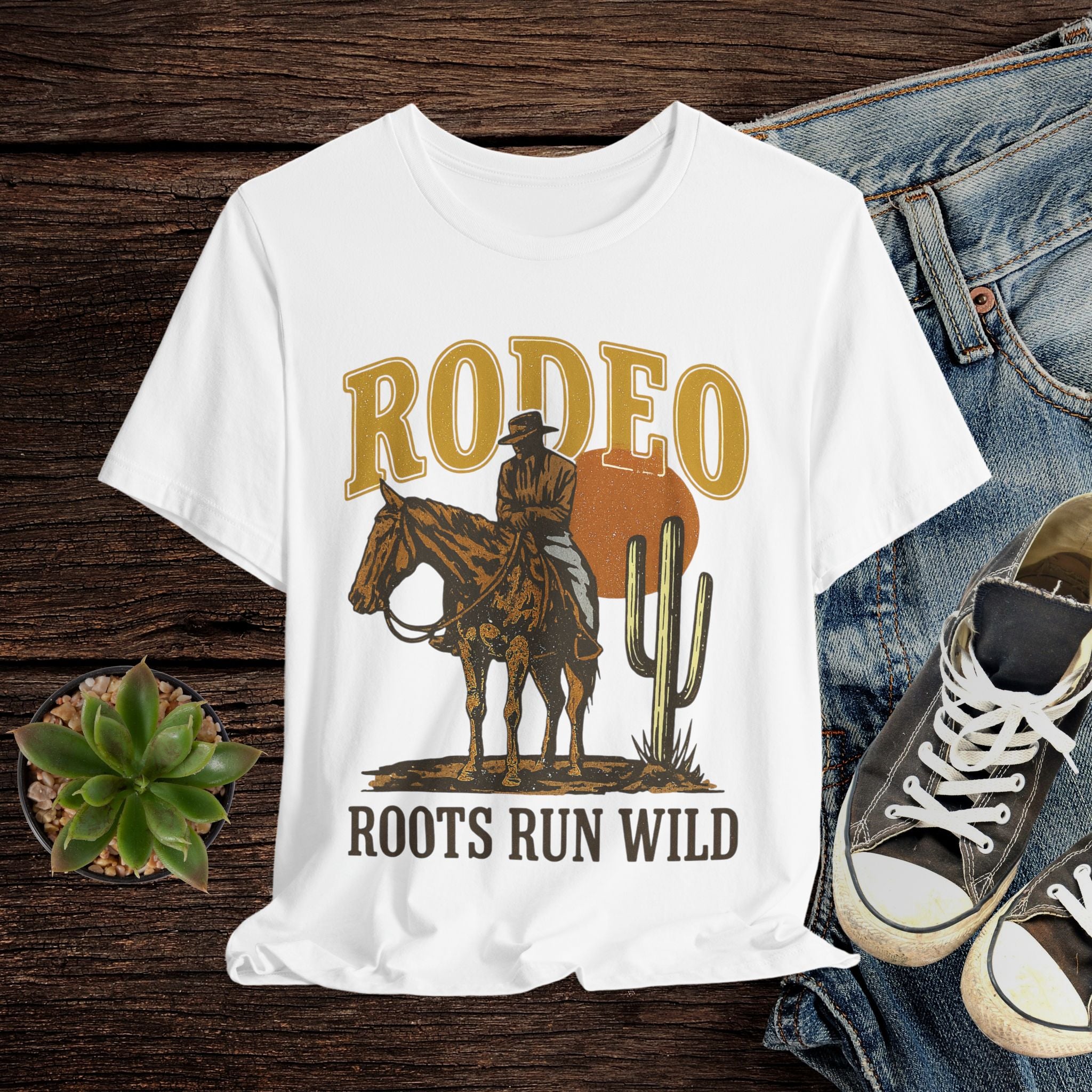 White t-shirt with a cowboy graphic and 'Rodeo Roots Run Wild' text on a wooden surface with jeans and shoes.