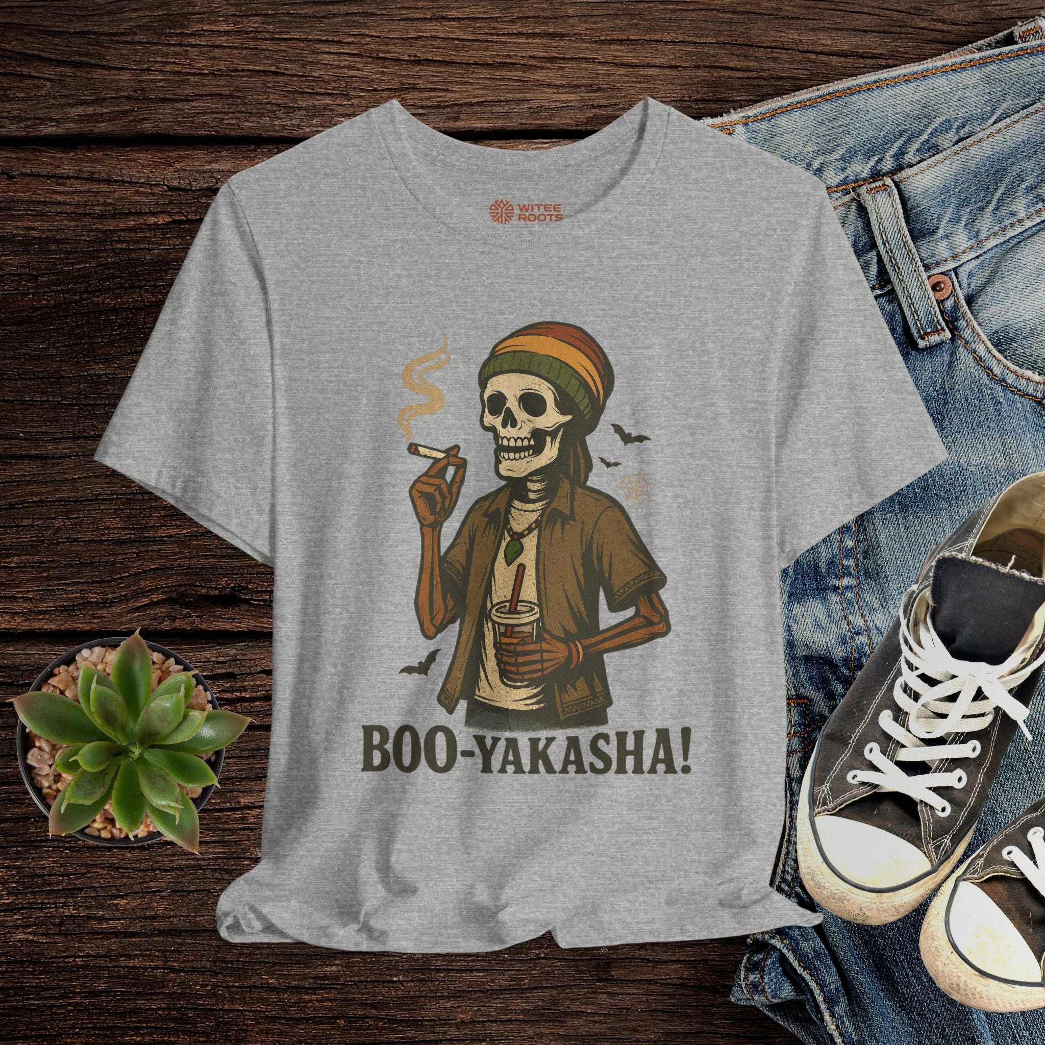 Gray t-shirt with a skeleton wearing a hat and smoking a cigarette, labeled 'BOO-YAKASHA!', displayed with jeans and sneakers on a wooden surface.