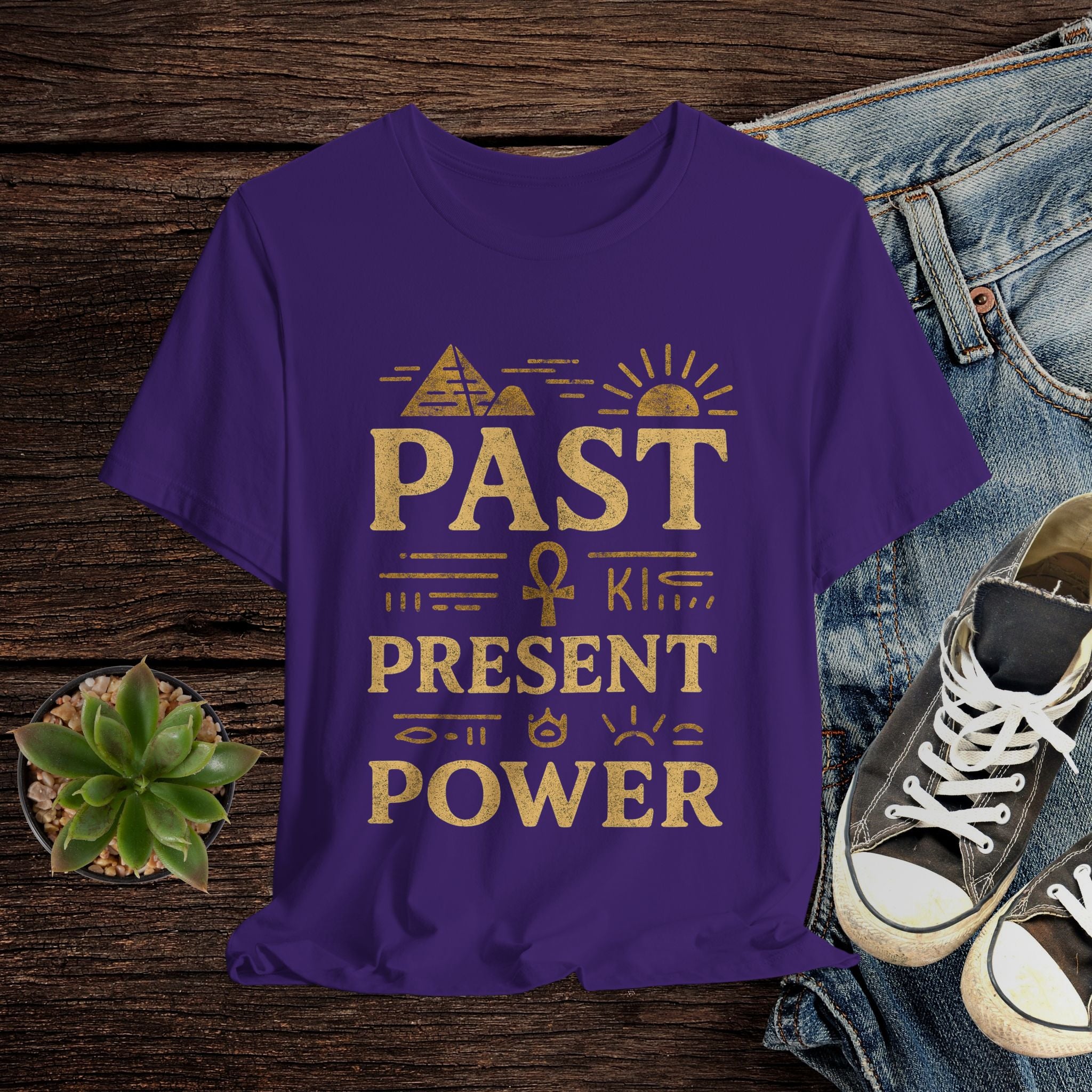Purple t-shirt with gold text on a wooden surface with jeans and sneakers.
