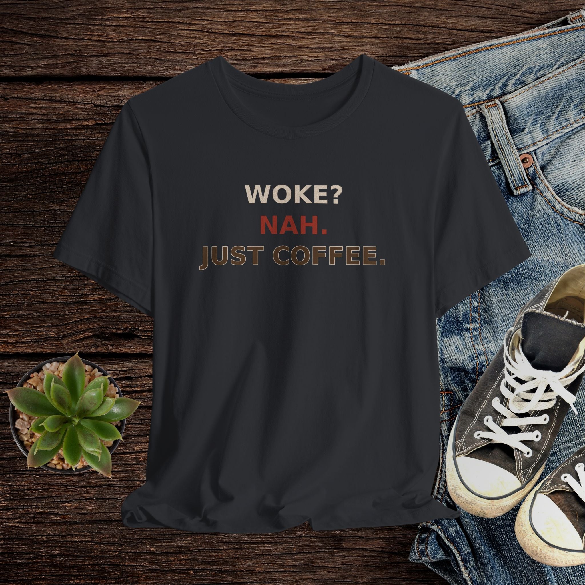 Black t-shirt with 'WOKE? NAH. JUST COFFEE.' text on a wooden surface with jeans and sneakers.