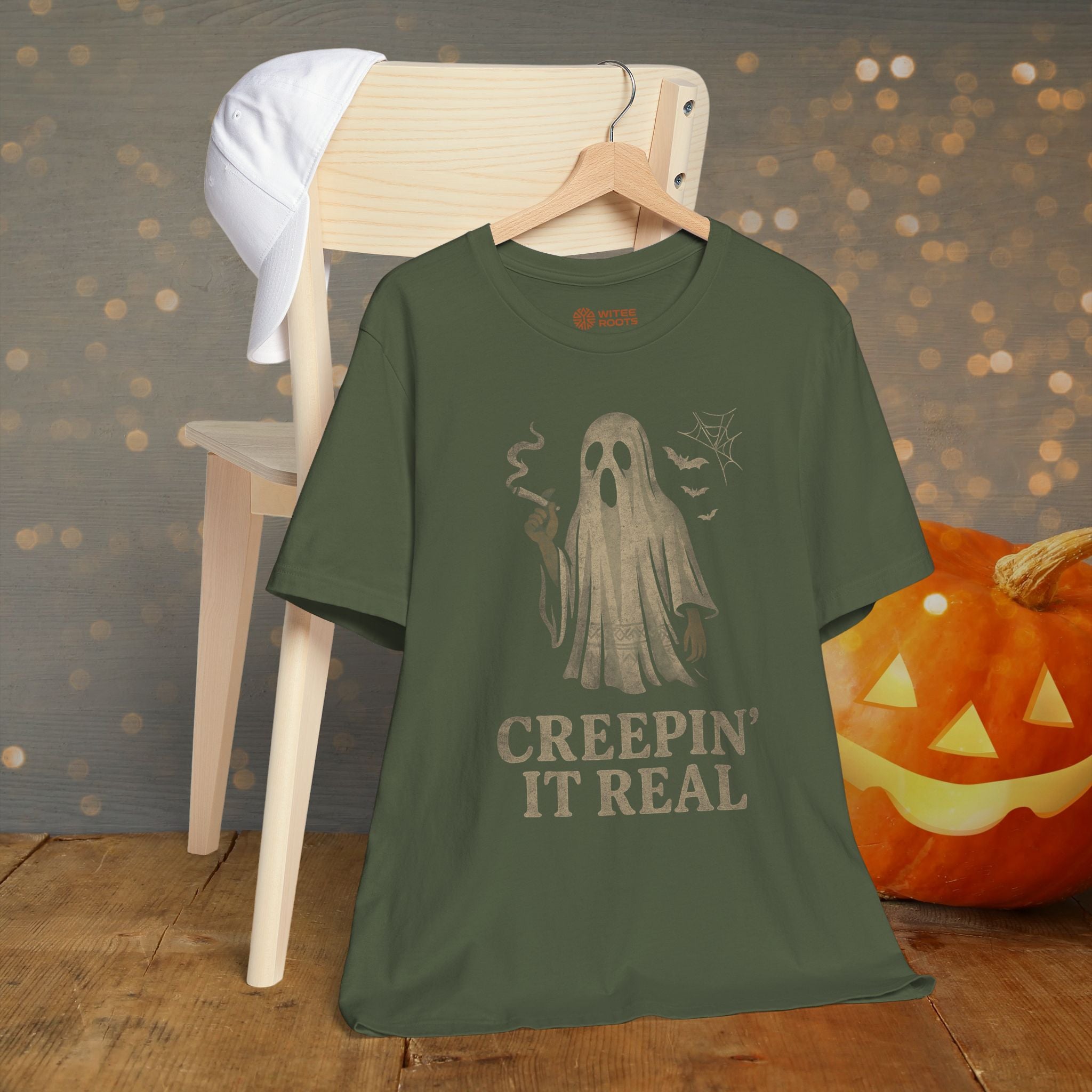 Green t-shirt with ghost graphic and text 'Creepin' It Real' on a wooden surface with a pumpkin and chair in the background.