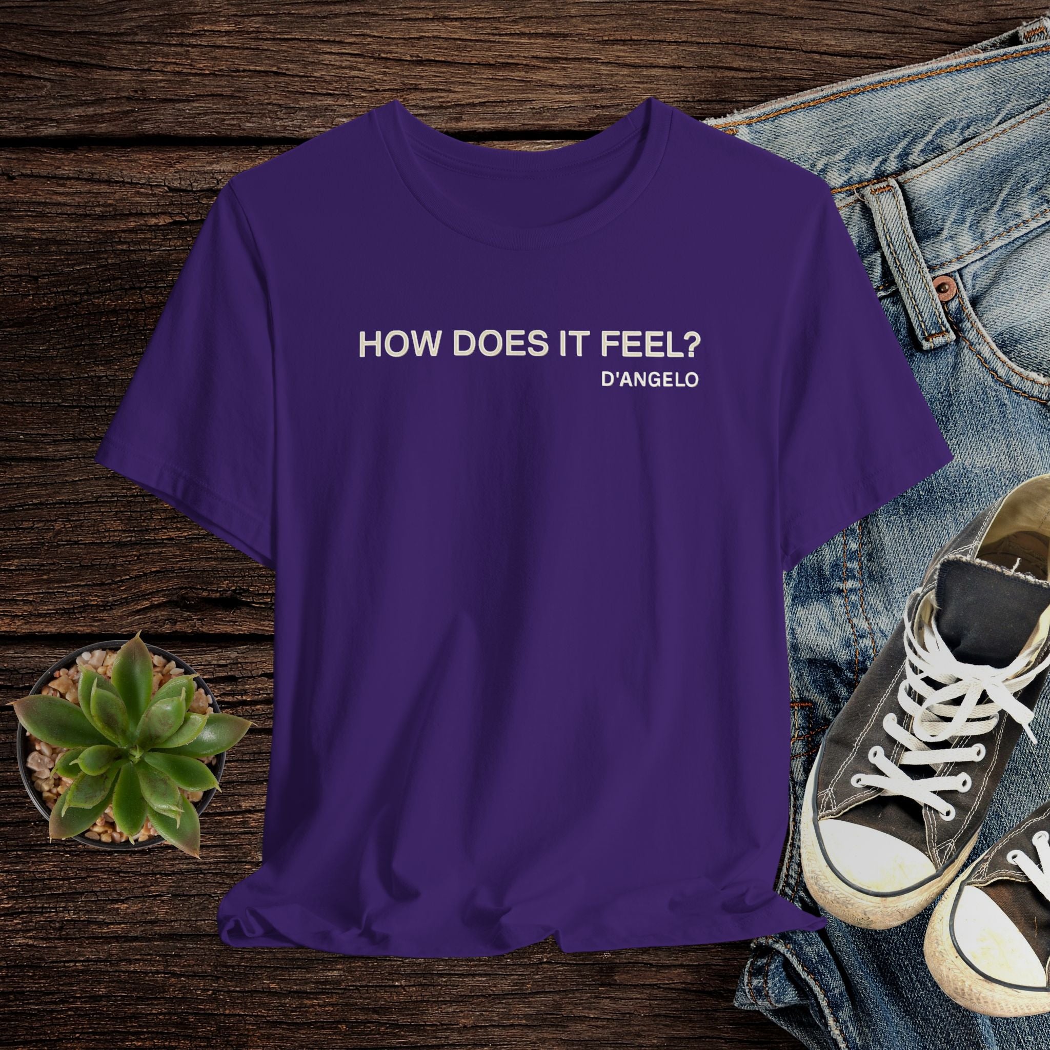 Purple t-shirt with text 'HOW DOES IT FEEL? D'ANGELO' on a wooden surface with jeans and sneakers.