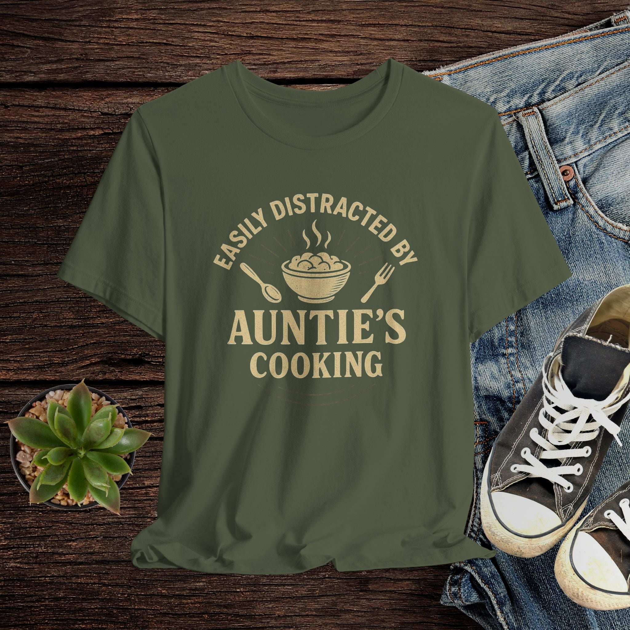 Green t-shirt with 'Easily Distracted by Auntie's Cooking' text on a wooden surface with jeans and shoes.
