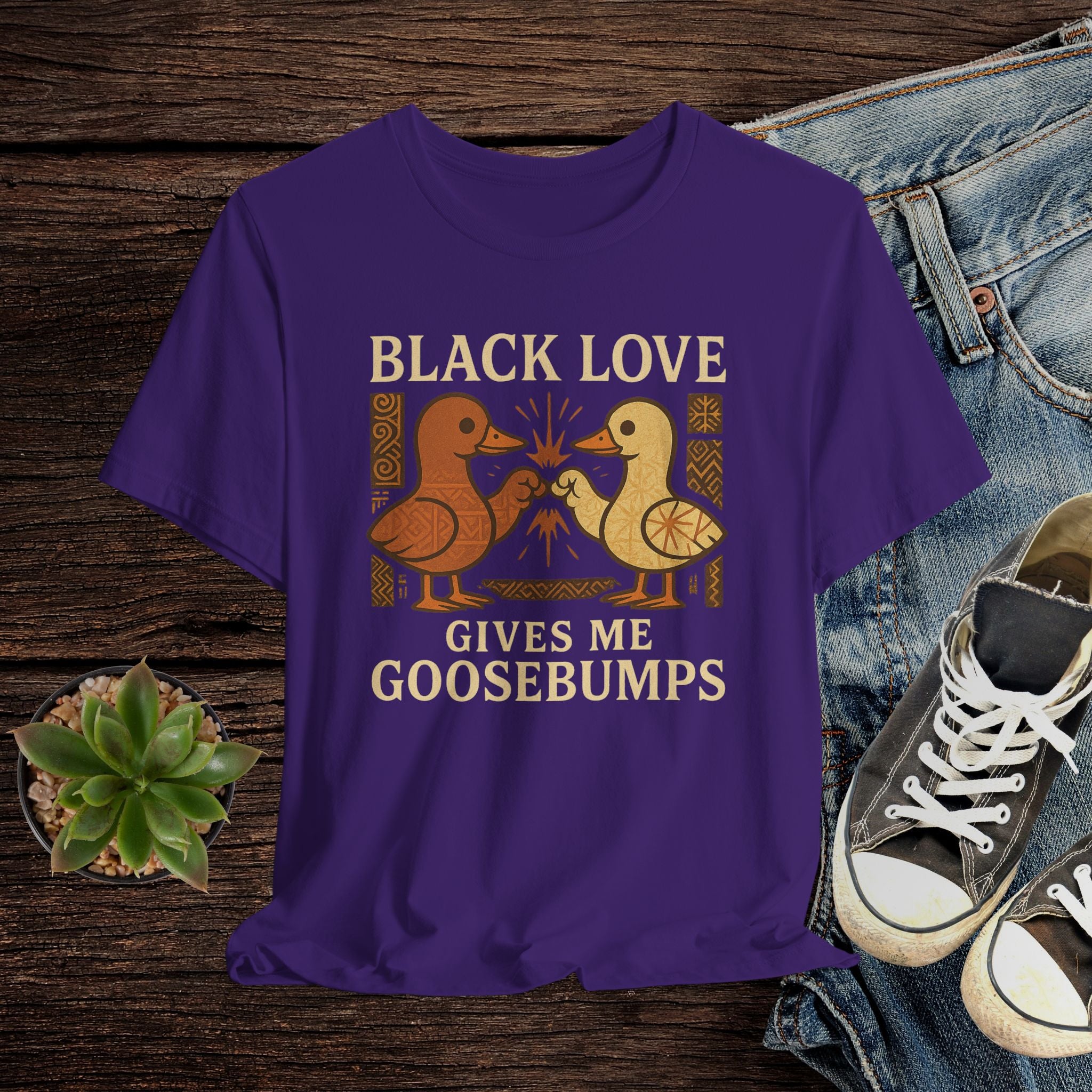 Purple t-shirt with 'Black Love Gives Me Goosebumps' text and graphic on a wooden surface with jeans and sneakers.