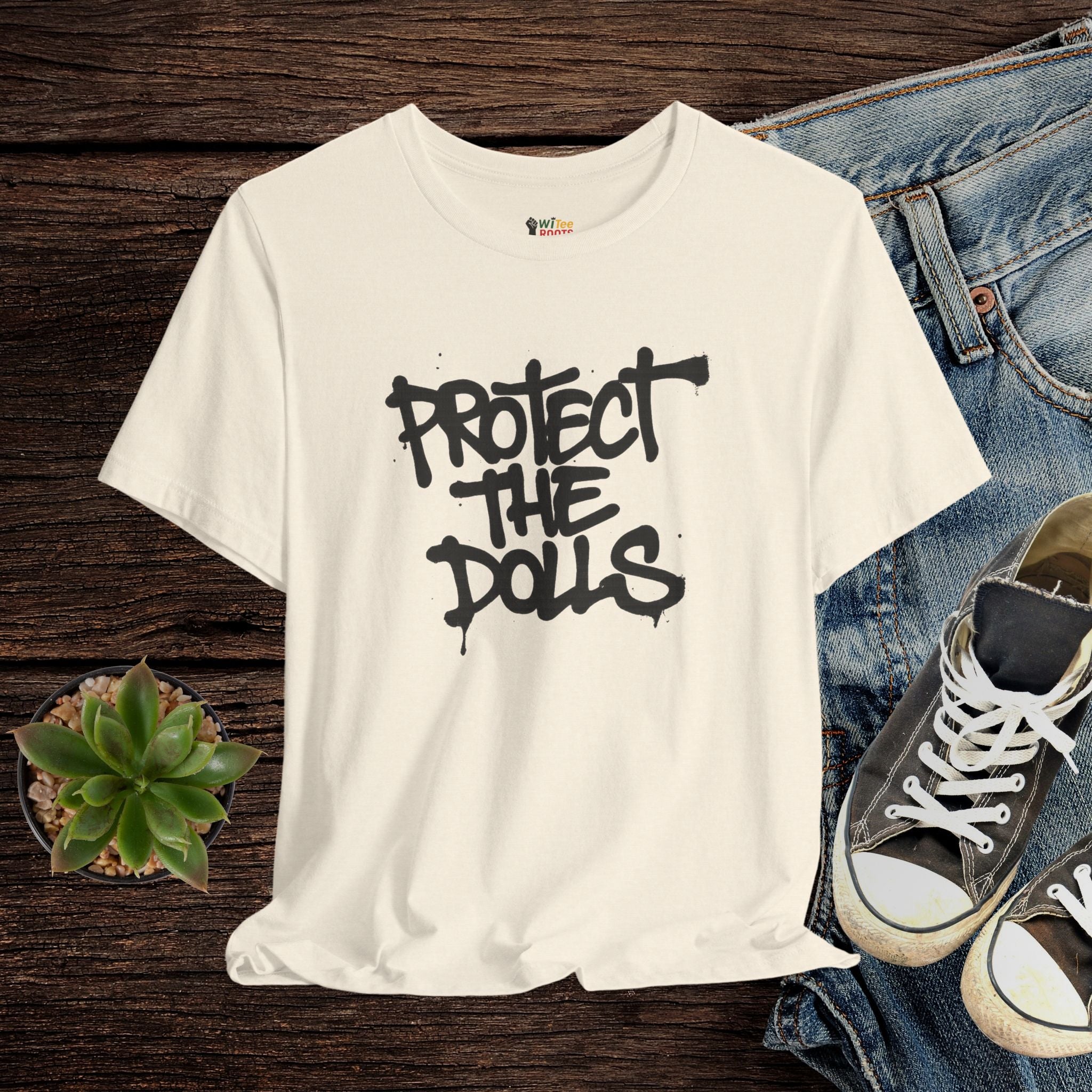 Beige t-shirt with 'PROTECT THE DOLLS' text on a wooden surface with jeans and sneakers.