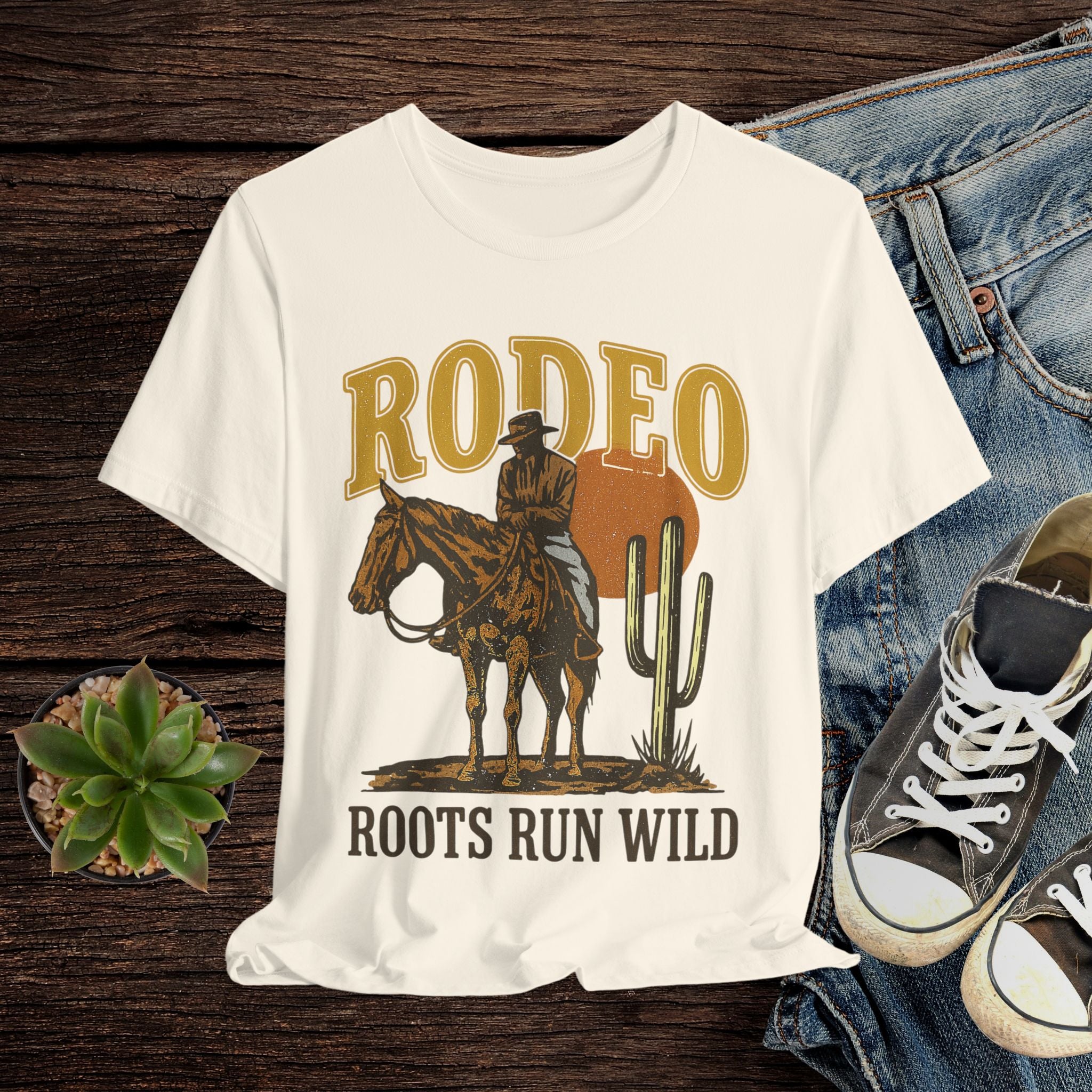 Cream t-shirt with a cowboy graphic and 'Rodeo Roots Run Wild' text on a wooden surface with jeans and shoes.