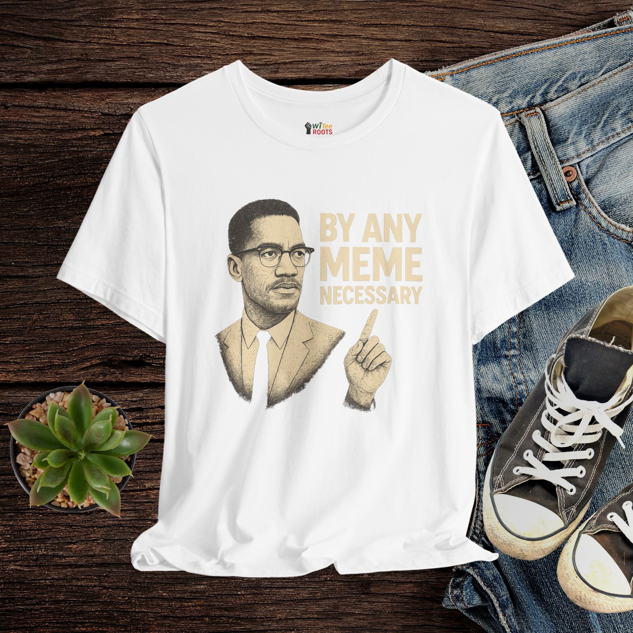 White t-shirt with a graphic of a man and text 'By Any Meme Necessary' on a wooden surface with jeans and shoes.