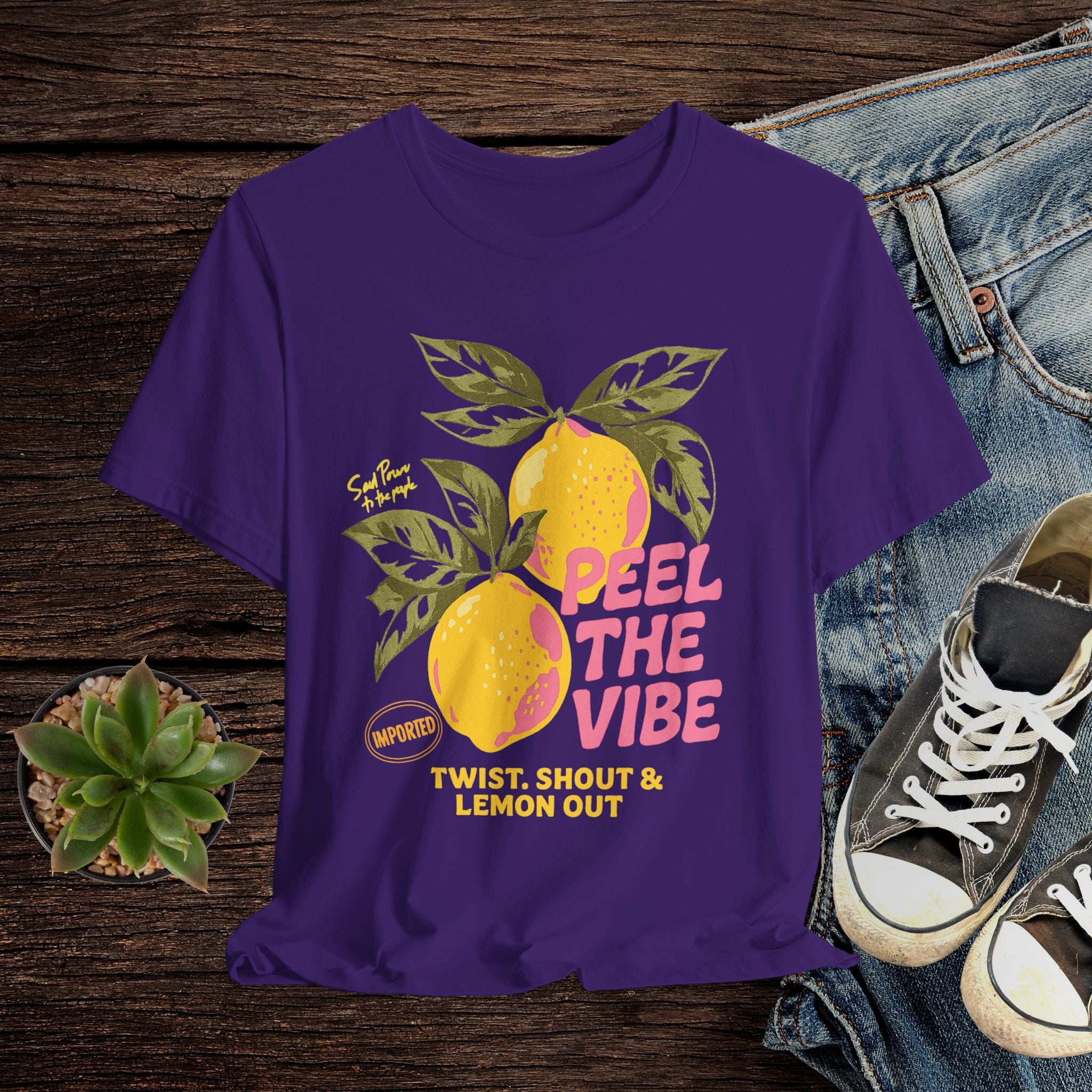 Purple t-shirt with lemon graphic and text on a wooden surface with jeans and sneakers.