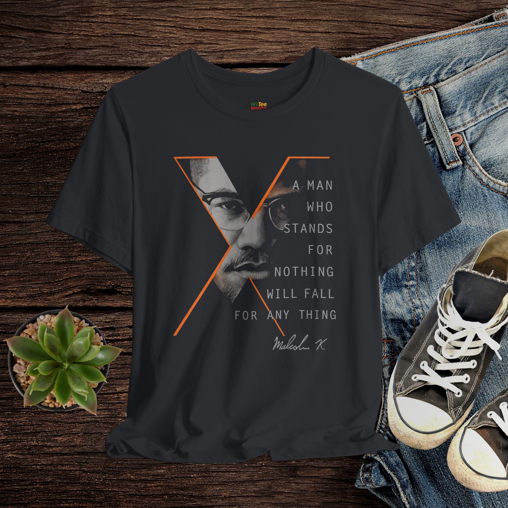 Black t-shirt with a graphic design and text on a wooden surface with jeans and sneakers.