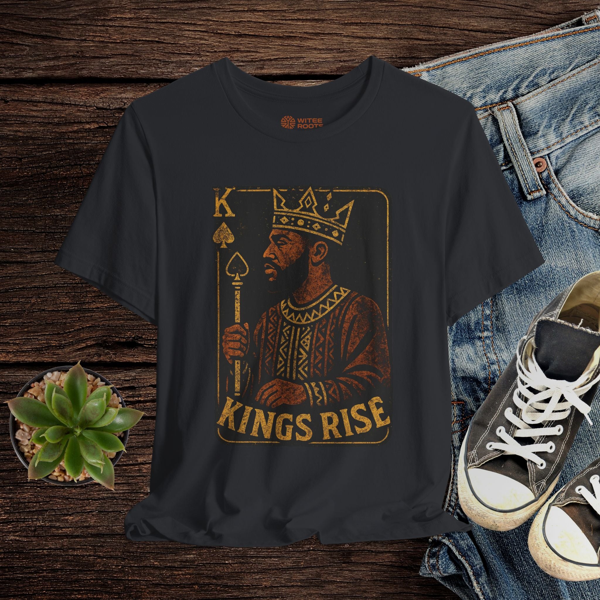 Black t-shirt with 'Kings Rise' graphic design on a wooden surface with jeans and sneakers.