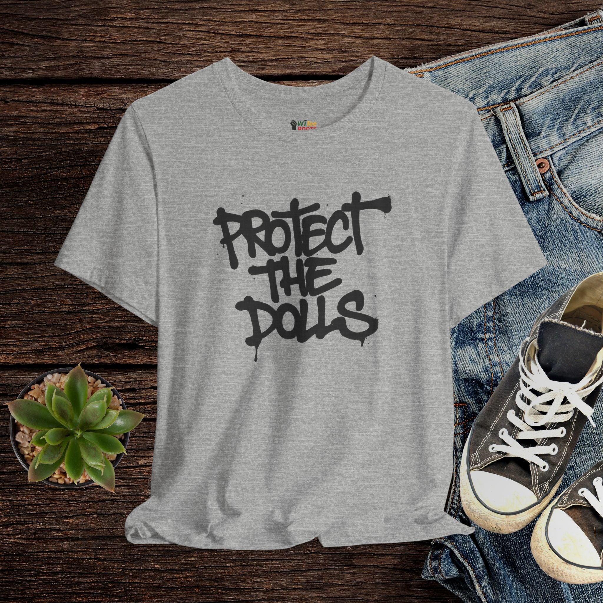 Gray t-shirt with 'PROTECT THE DOLLS' text, paired with jeans and sneakers on a wooden surface.