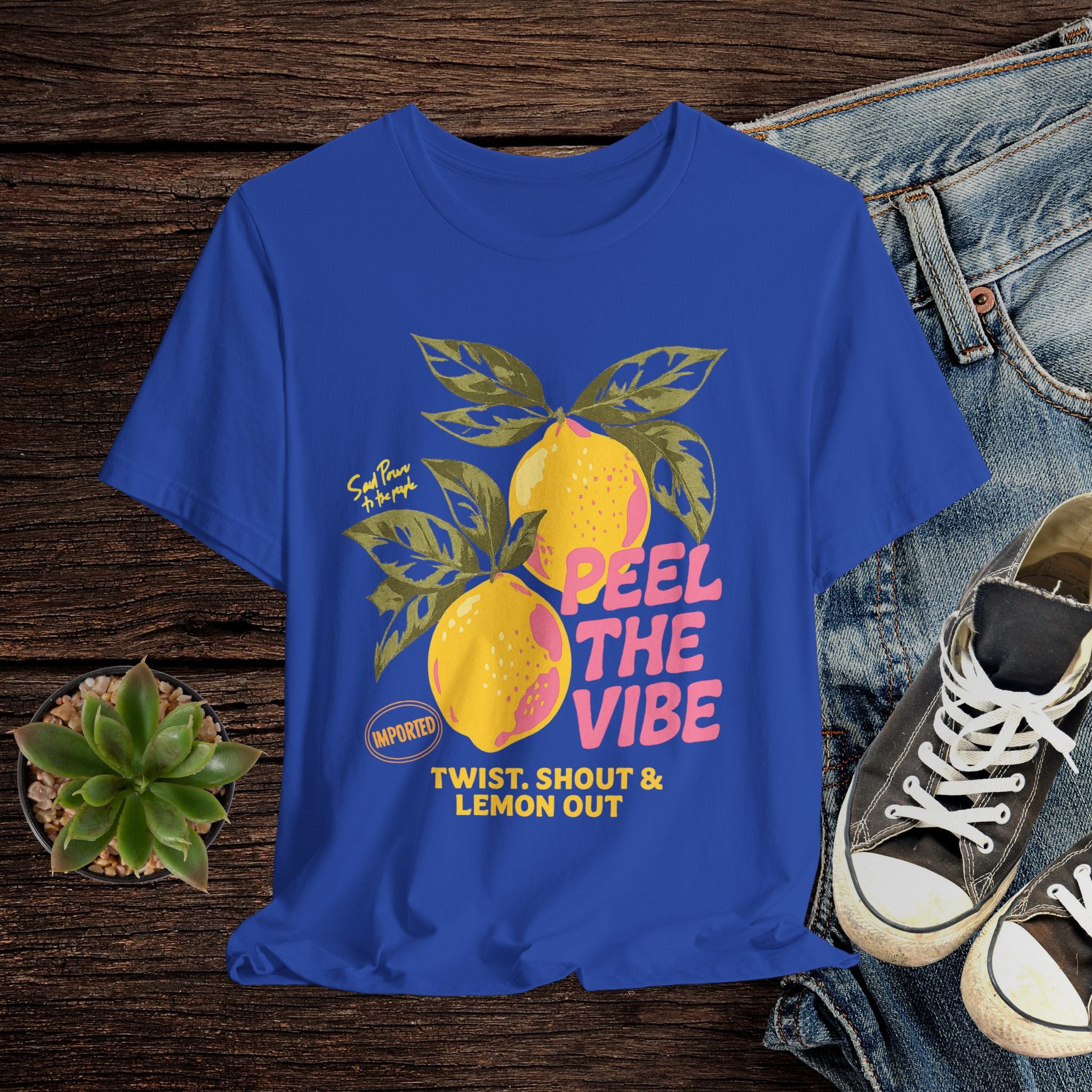 Blue t-shirt with lemon graphic and text on a wooden surface with jeans and sneakers.