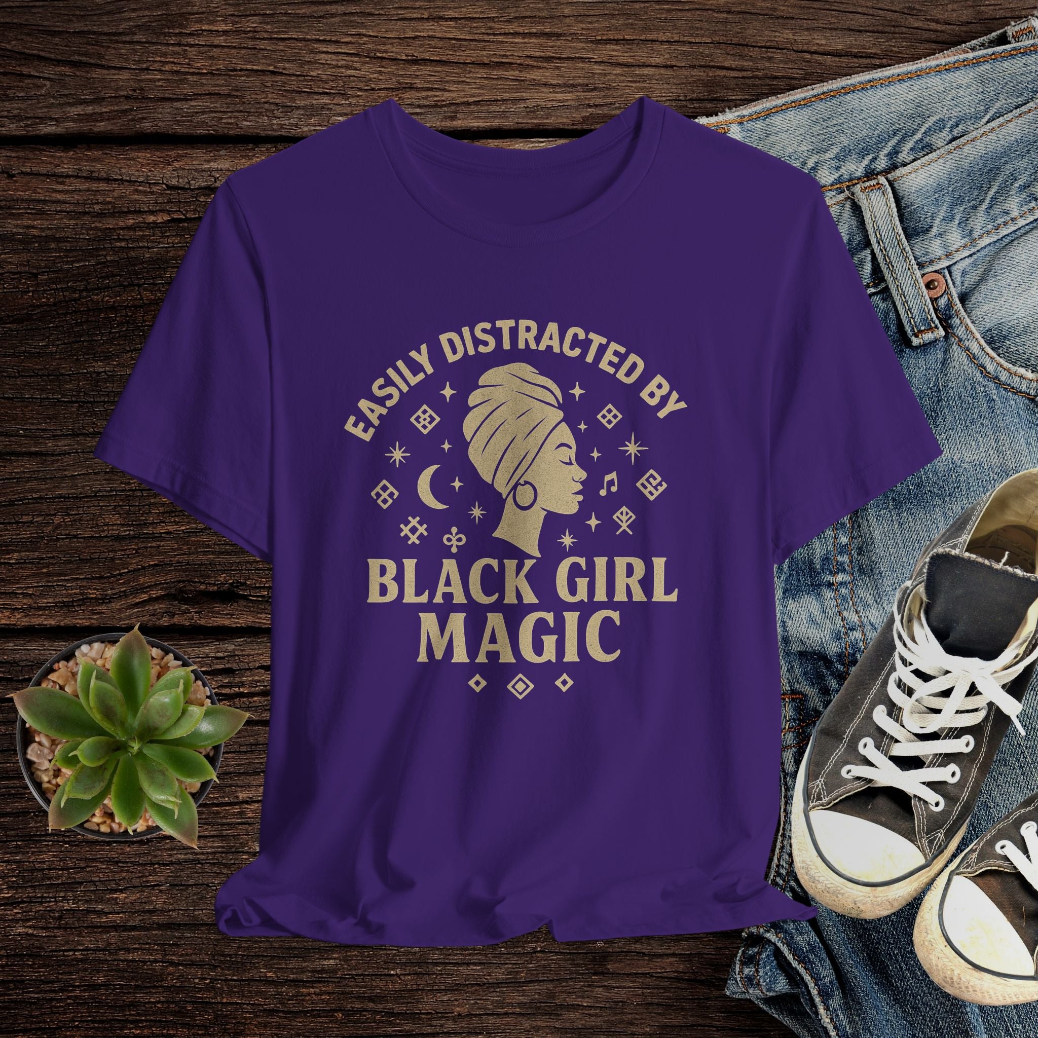 Purple t-shirt with 'Easily Distracted by Black Girl Magic' design on a wooden surface with jeans and sneakers.