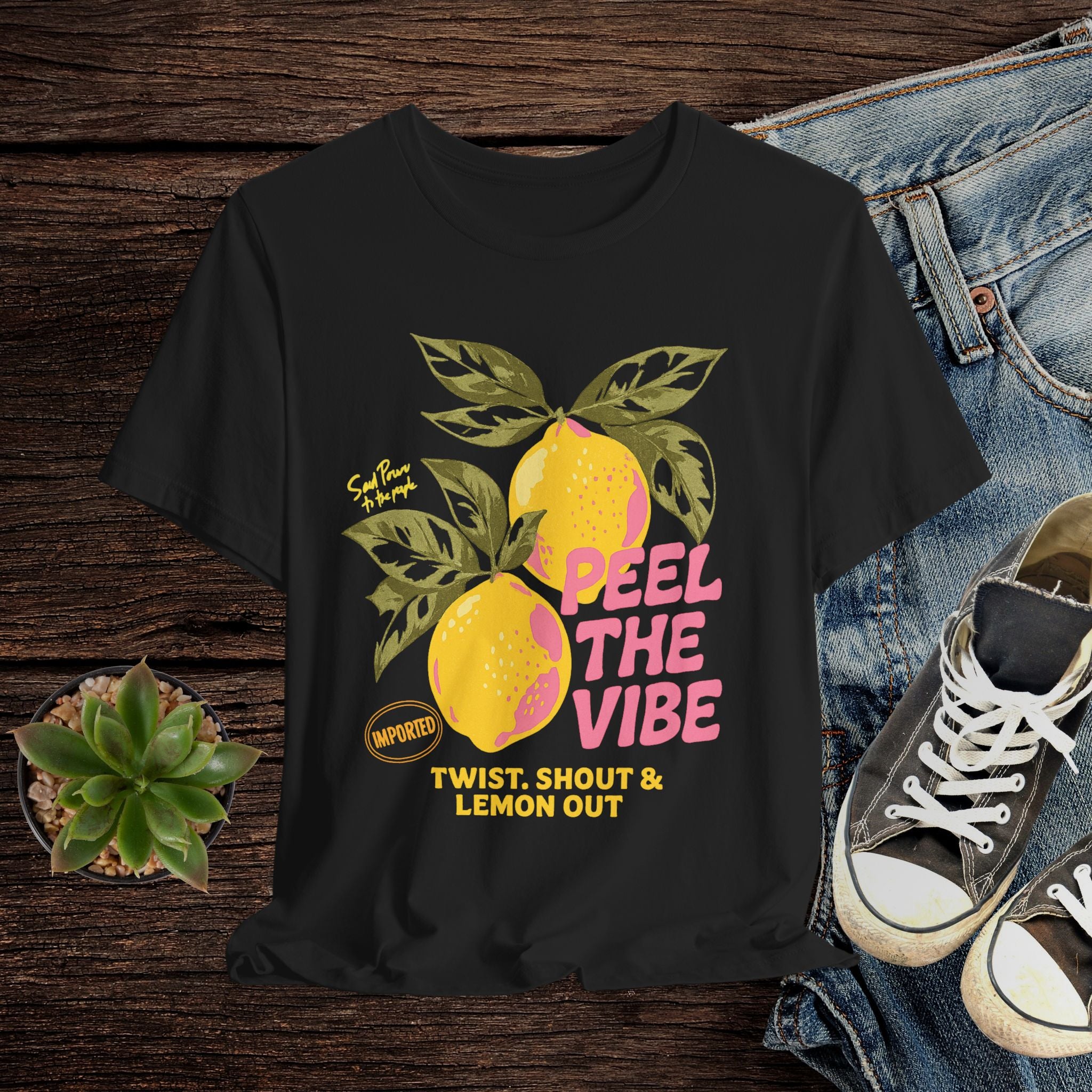 Black t-shirt with lemon graphic and text on a wooden surface with jeans and sneakers.