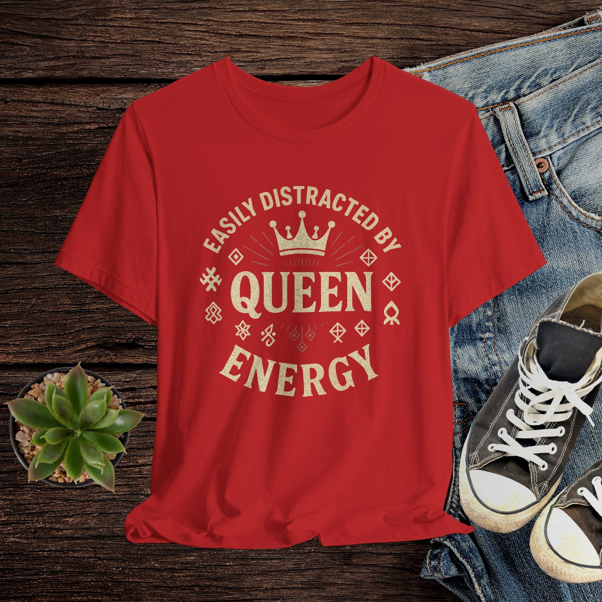 Red t-shirt with 'Easily Distracted by Queen Energy' text on a wooden surface with jeans and sneakers.