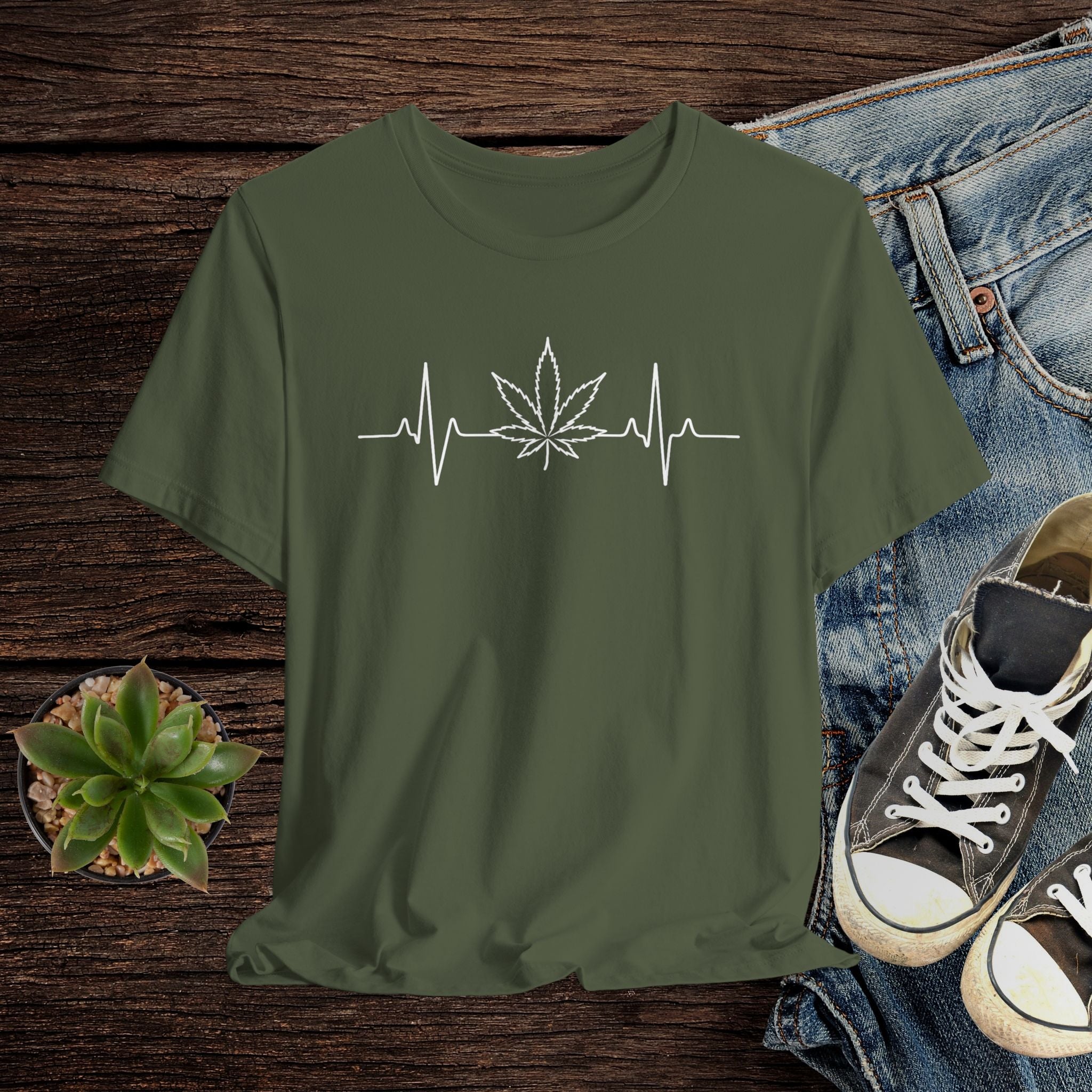 Green t-shirt with a cannabis leaf and heartbeat line design on a wooden surface with jeans and shoes.