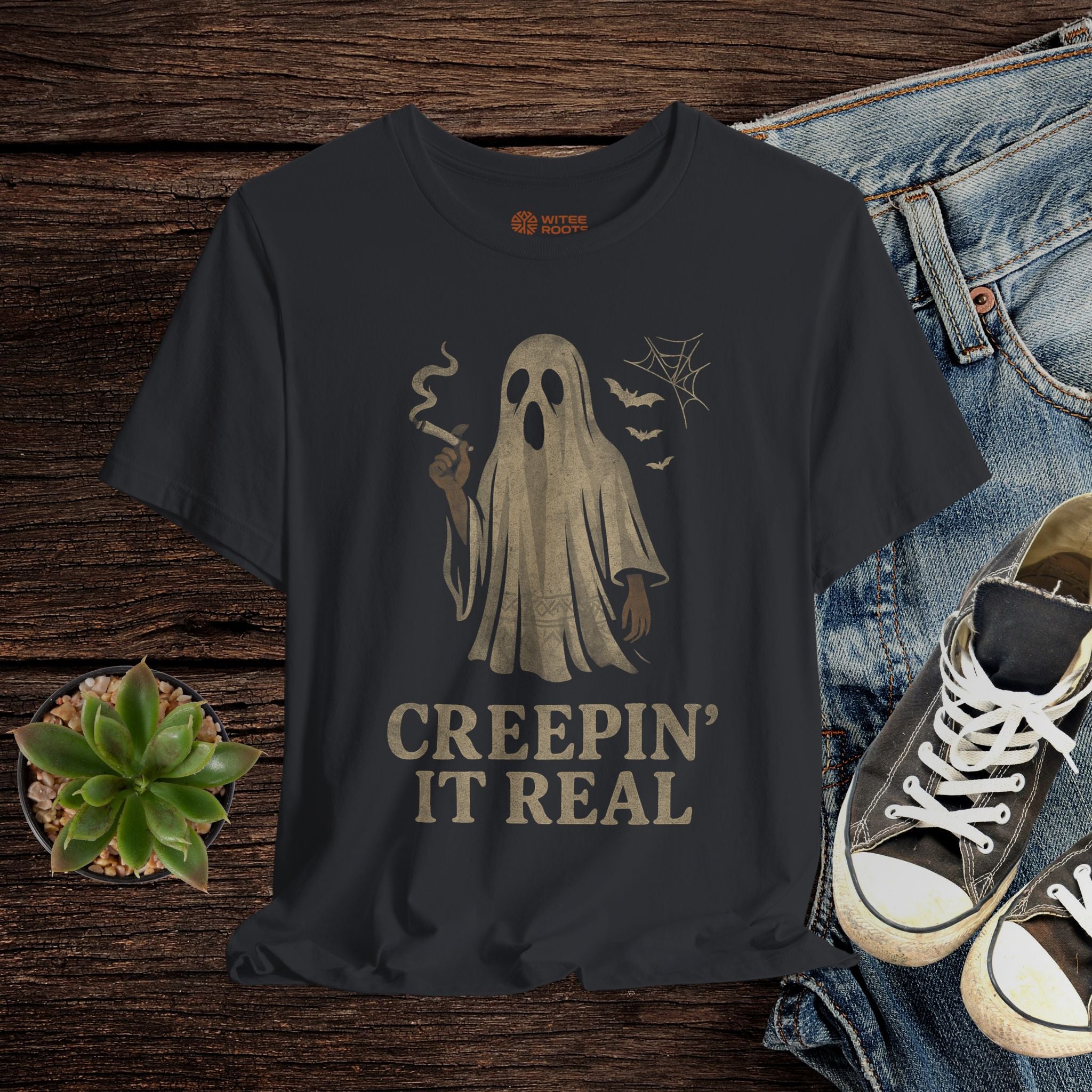 Black t-shirt with ghost graphic and 'Creepin' It Real' text on a wooden surface with jeans and sneakers.
