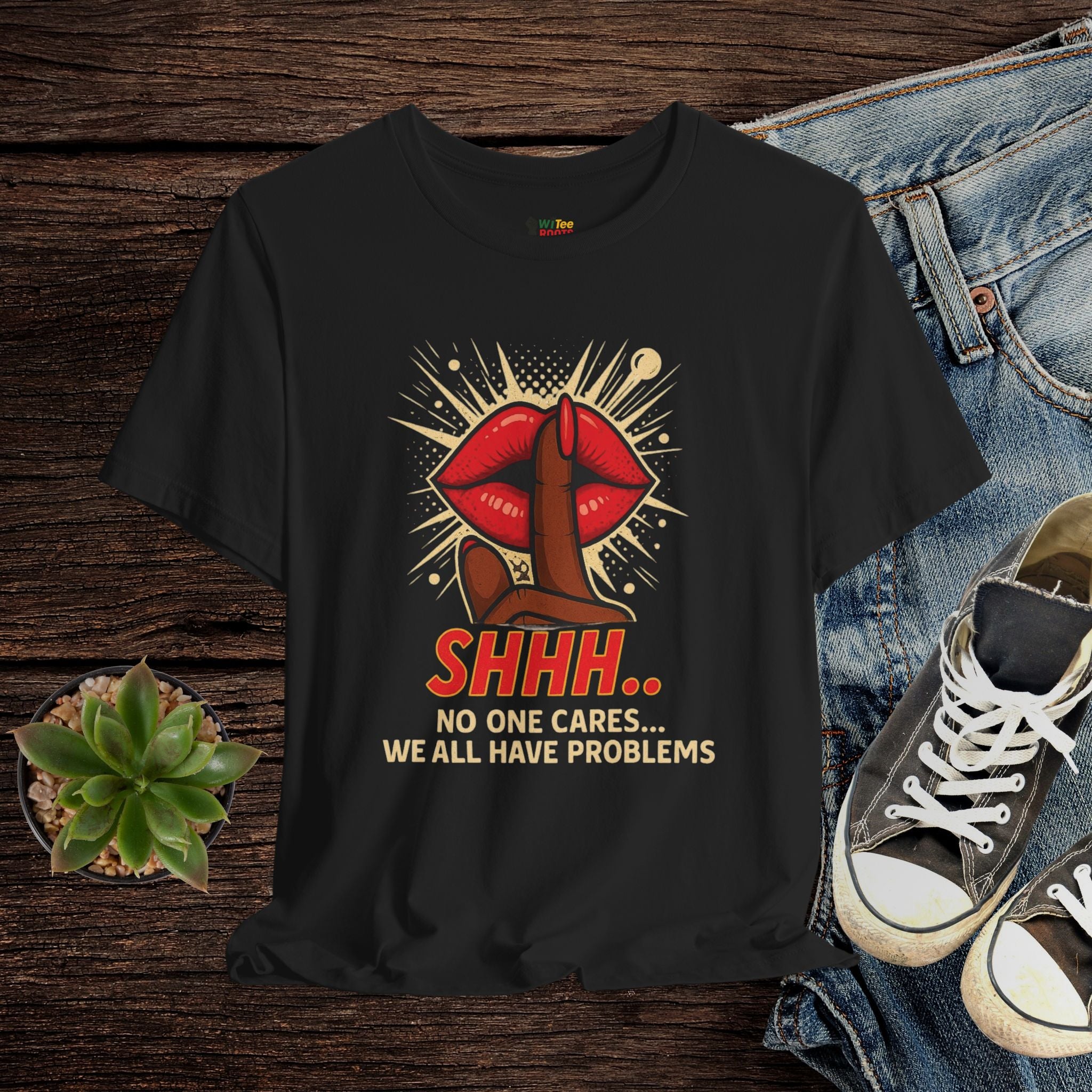 Black t-shirt with graphic design and text on a wooden surface with jeans and sneakers.