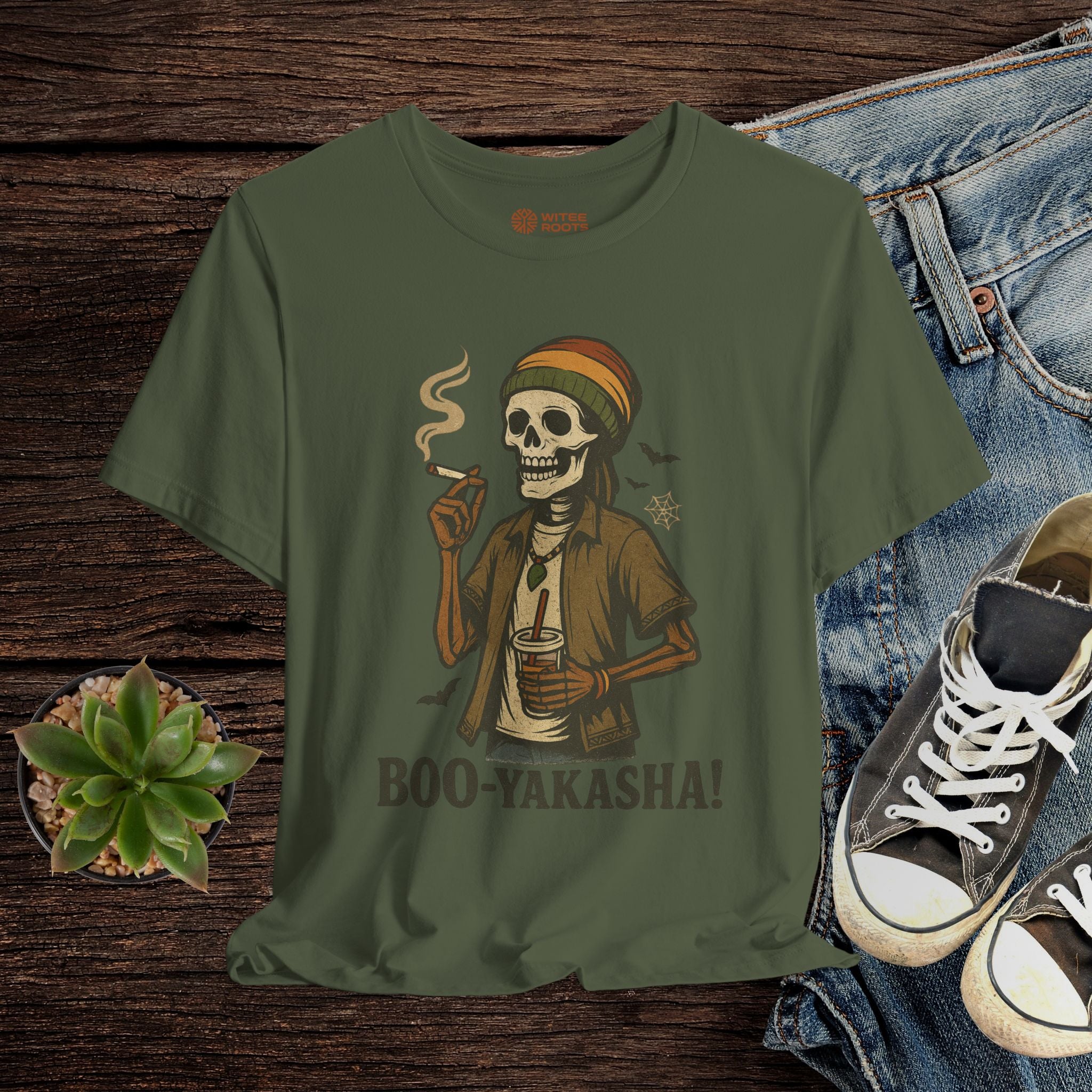 Green t-shirt with a skeleton wearing a beanie and smoking a cigarette, placed on a wooden surface with jeans and sneakers.