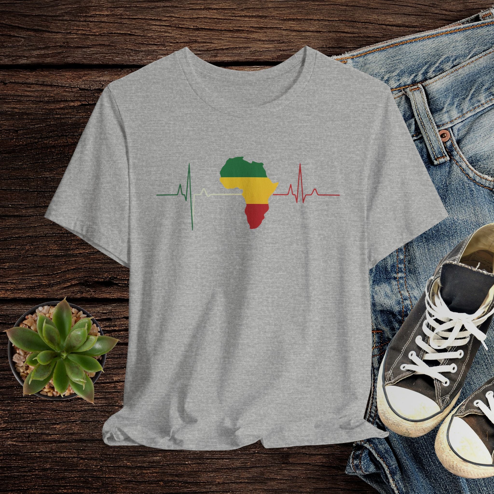 Gray t-shirt with a heart rate design featuring Africa and the colors of the Pan-African flag, displayed on a wooden surface with jeans and shoes.