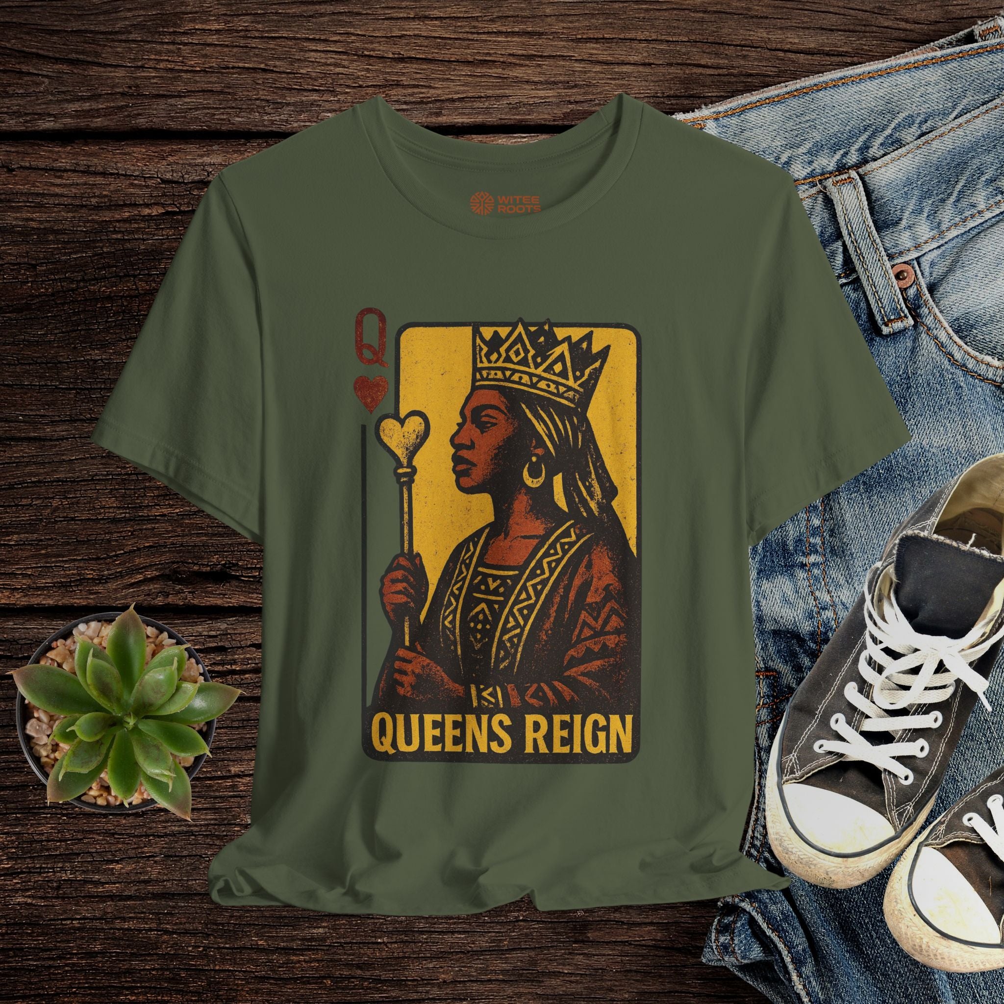 Green t-shirt with 'Queens Reign' design on a wooden surface with jeans and sneakers.