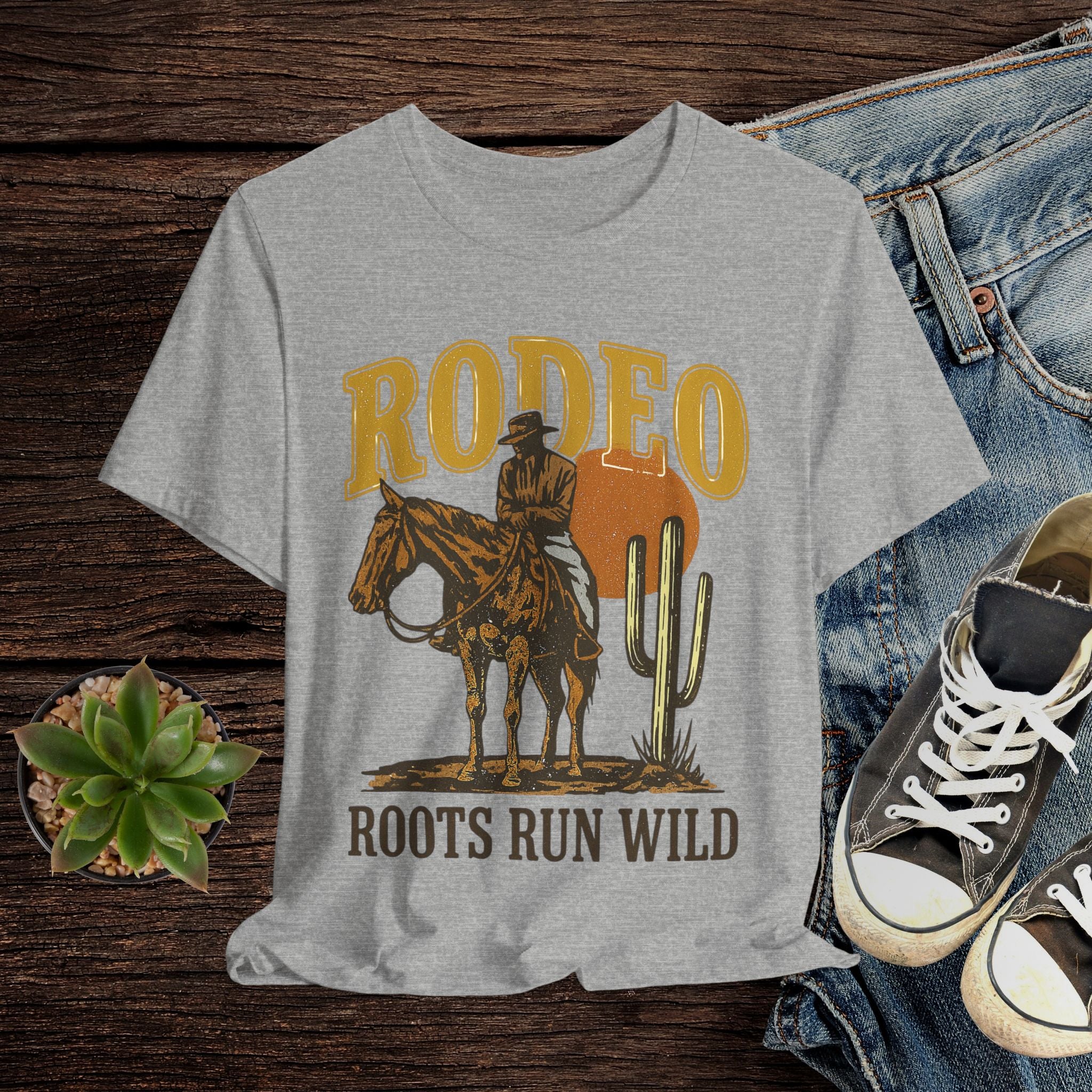 Gray t-shirt with a cowboy graphic and 'Rodeo Roots Run Wild' text on a wooden surface with jeans and shoes.