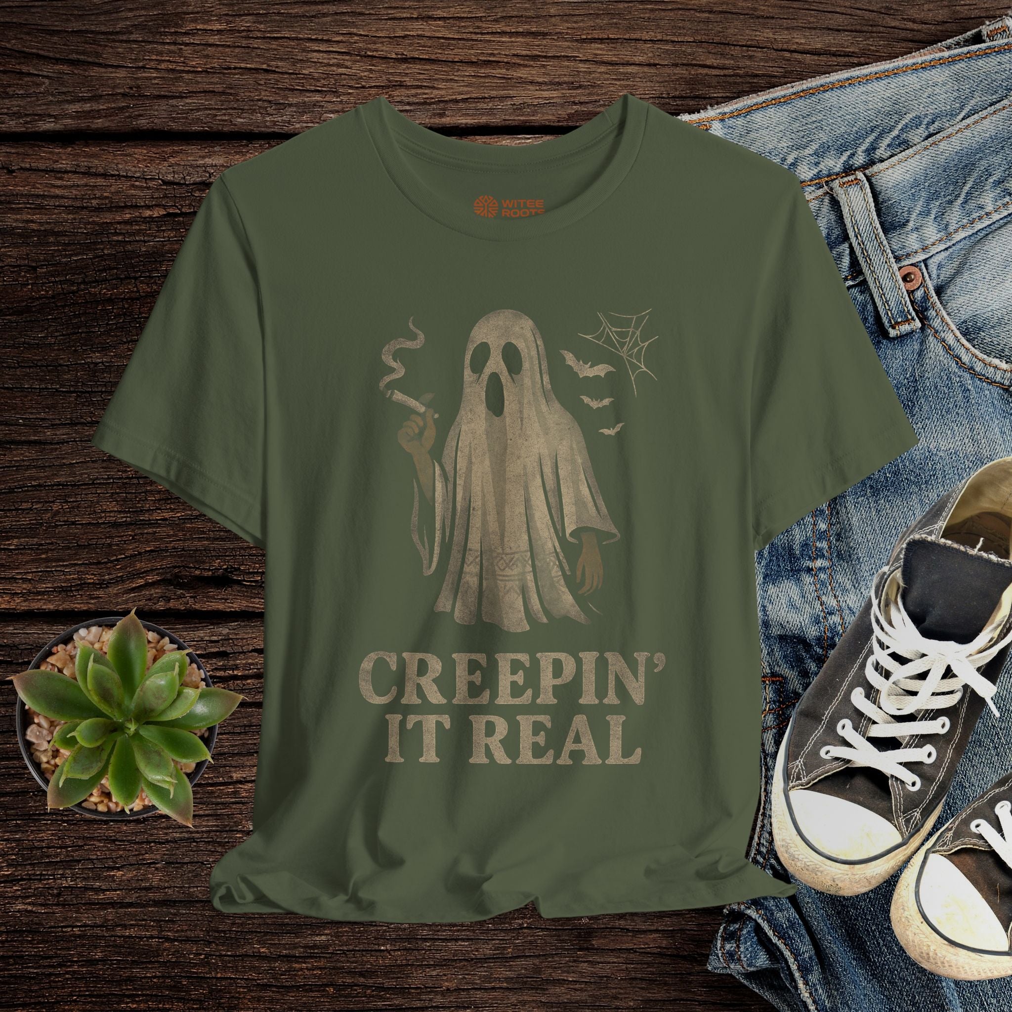 Green t-shirt with ghost graphic and 'Creepin' It Real' text on a wooden surface with jeans and sneakers.