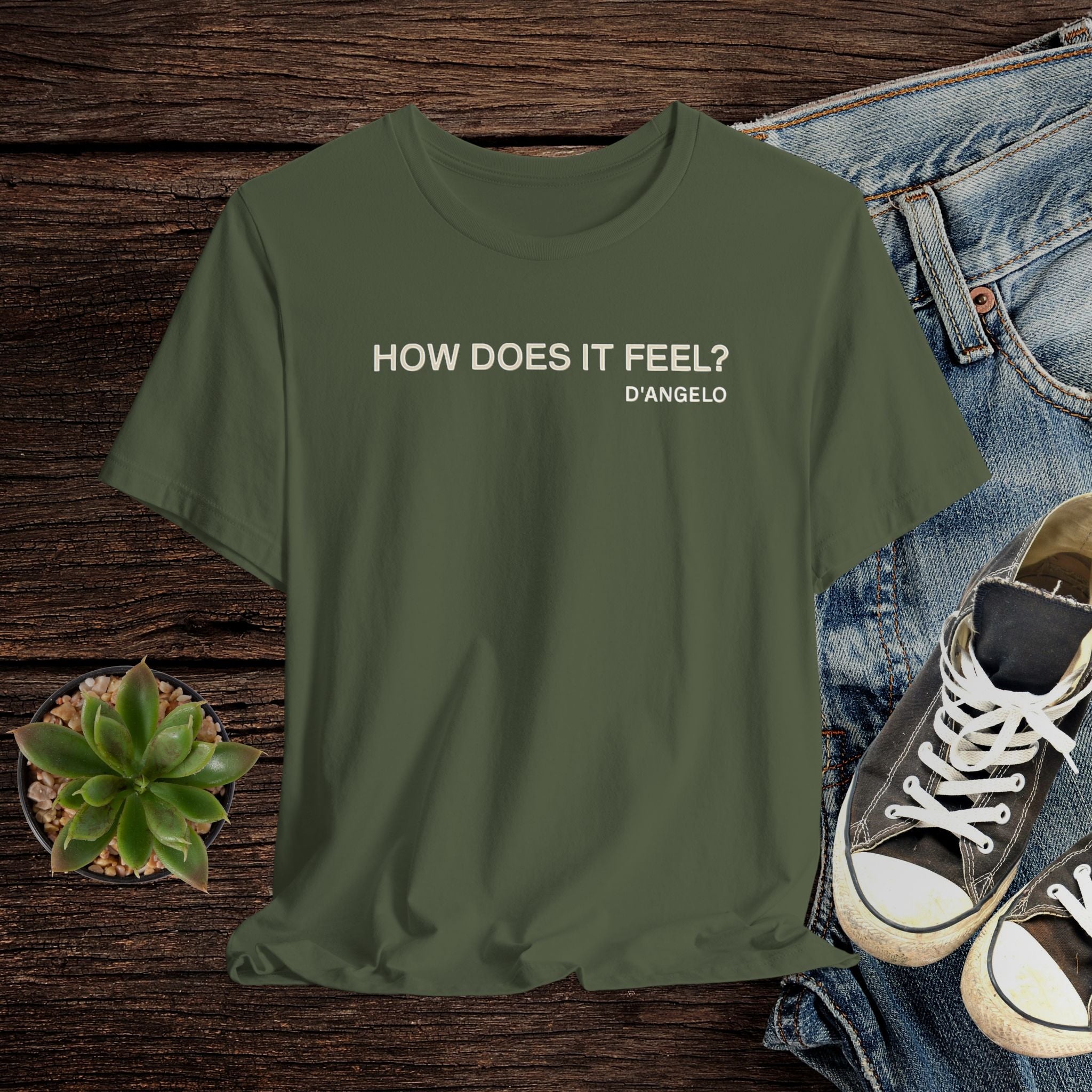 Green t-shirt with 'HOW DOES IT FEEL? D'ANGELO' text on a wooden surface with jeans and sneakers.