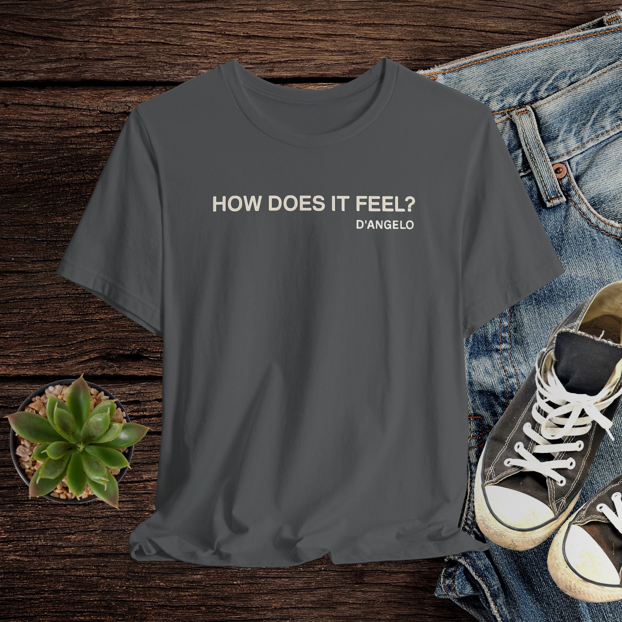 Gray t-shirt with 'HOW DOES IT FEEL? D'ANGELO' text on a wooden surface with jeans and sneakers.