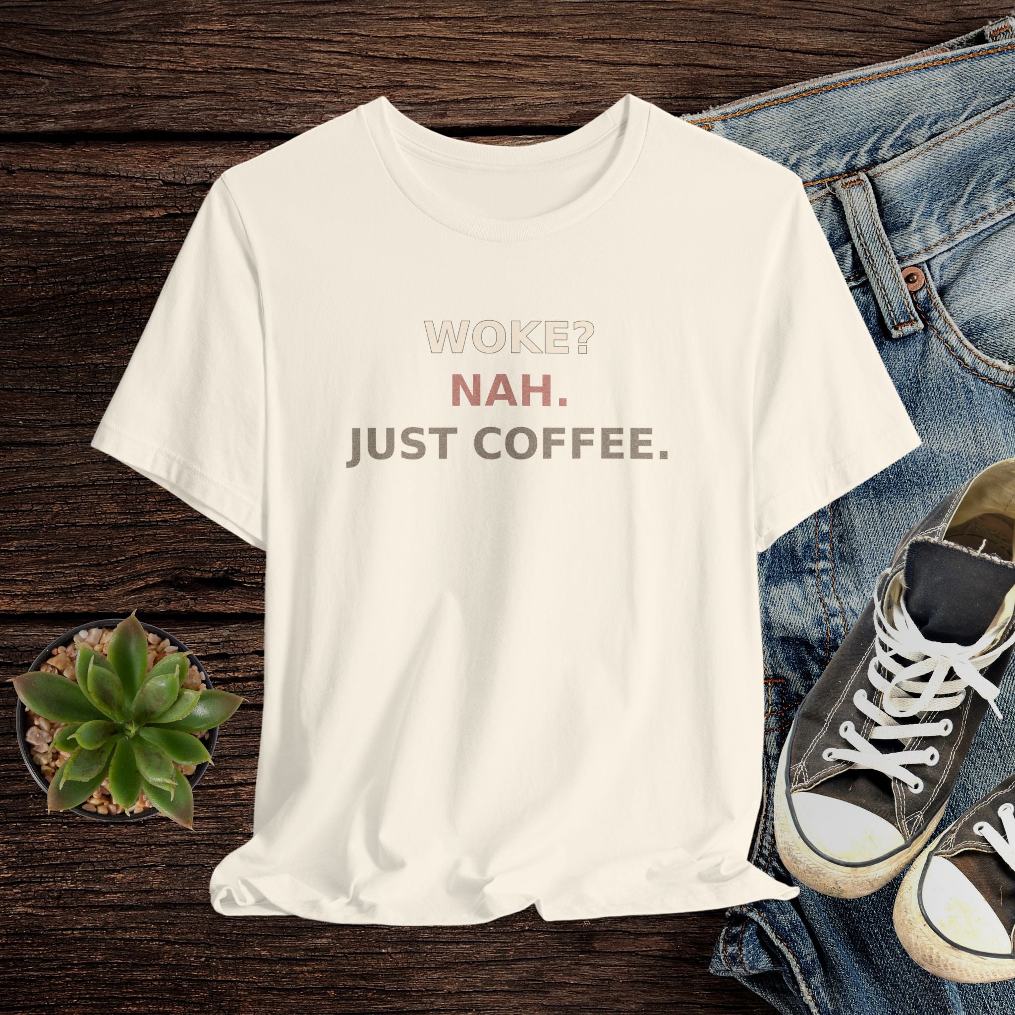 Beige t-shirt with 'Woke? Nah. Just Coffee.' text, paired with jeans and sneakers on a wooden surface.