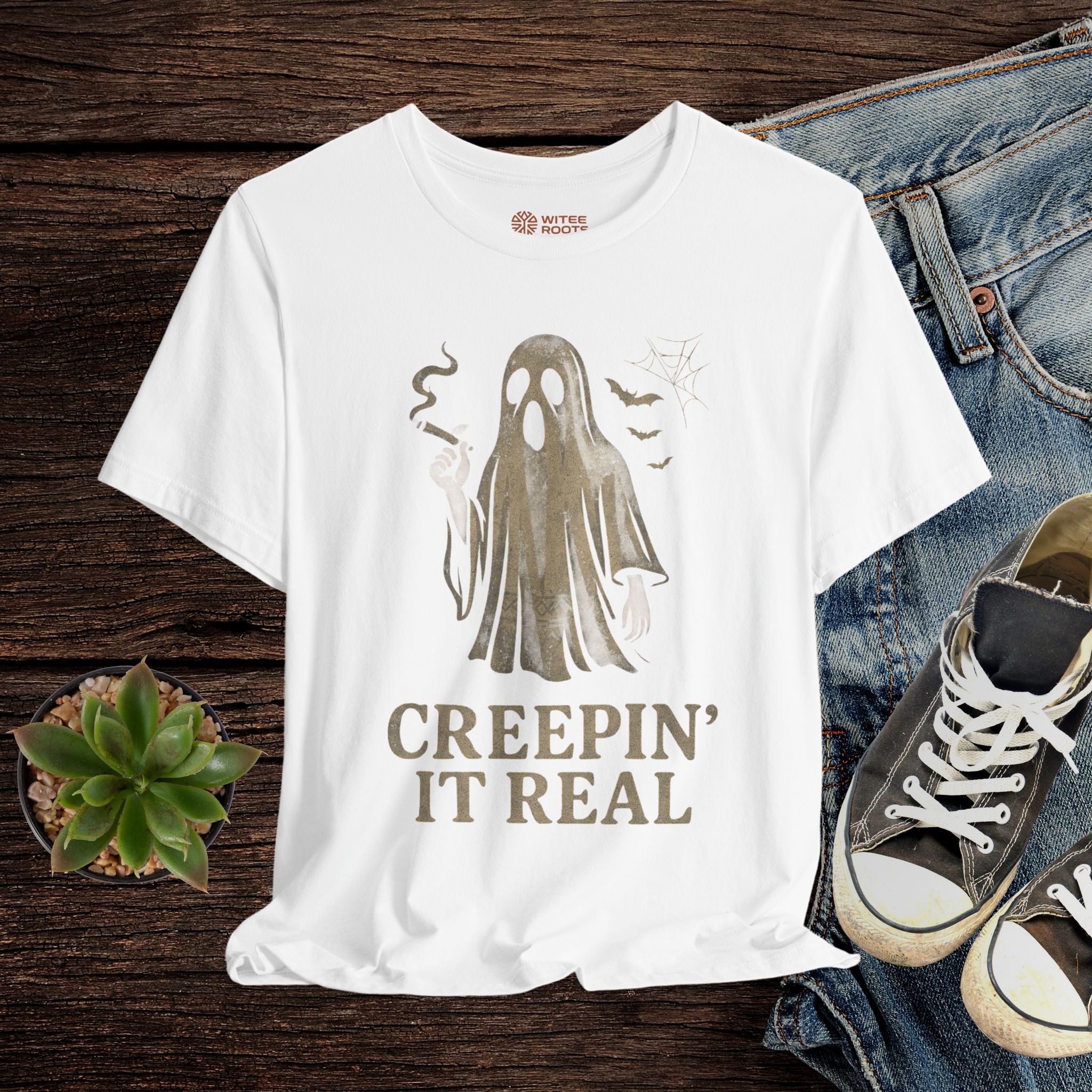 White t-shirt with ghost graphic and 'Creepin' It Real' text on a wooden surface with jeans and sneakers.